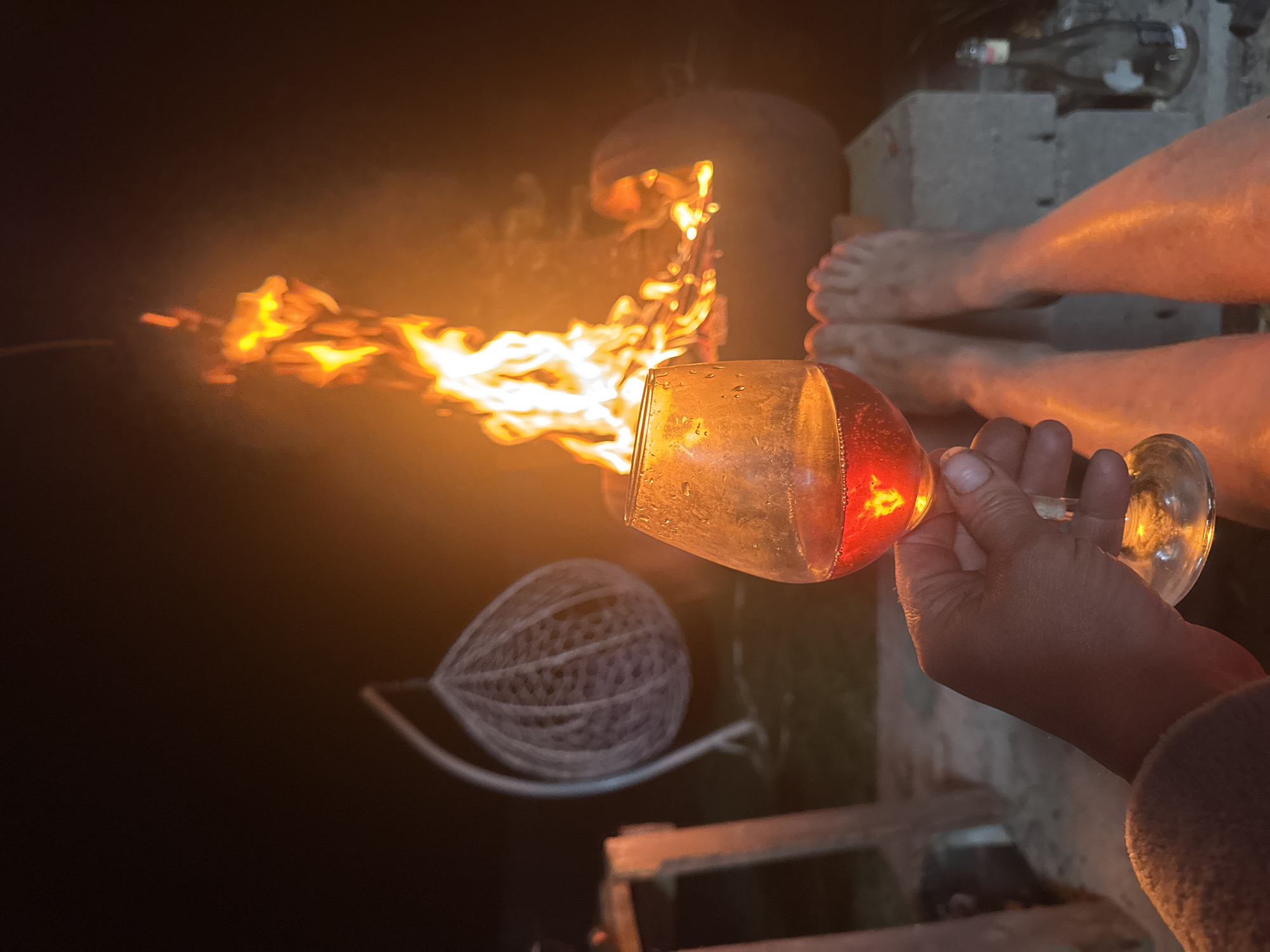 The image shows a hand holding a wine glass, partially filled with a red liquid, in front of a fire. The fire is intense and bright orange, emanating from a structure with a rounded top. A wicker ball is also visible in the background. Two pairs of bare feet can be seen next to the fire structure. An empty bottle lays on some concrete blocks. The scene appears to be outdoors, possibly in the evening or at night. There are no immediately visible clues within the image to confirm the location in Chaikovsky, Russia.