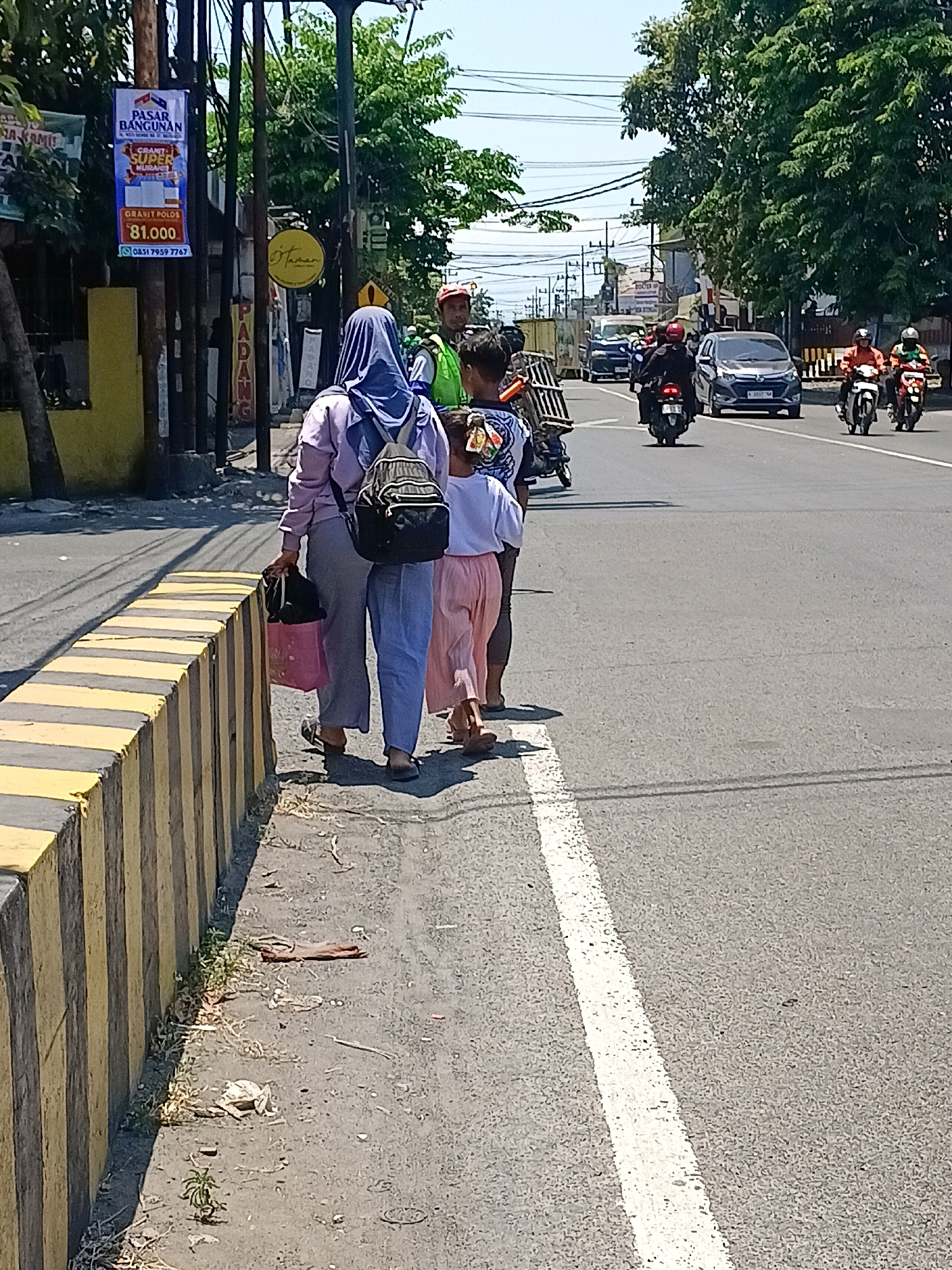 A street scene in Kalijaten, Indonesia, features three pedestrians, two women and one child, walking on the sidewalk beside a concrete barrier painted with yellow and black stripes. The woman in front wears a purple hijab and a light purple jacket over light blue pants, carrying a pink plastic bag in her right hand and a black backpack on her back. Beside her, a child in a white shirt and pink pants walks, followed closely by another person. In the background, traffic moves along a road, with several motorcycles and cars visible. A vendor with a red hat and green vest is also present near a utility pole adorned with banners. Trees line the street, and the sky is clear and blue.