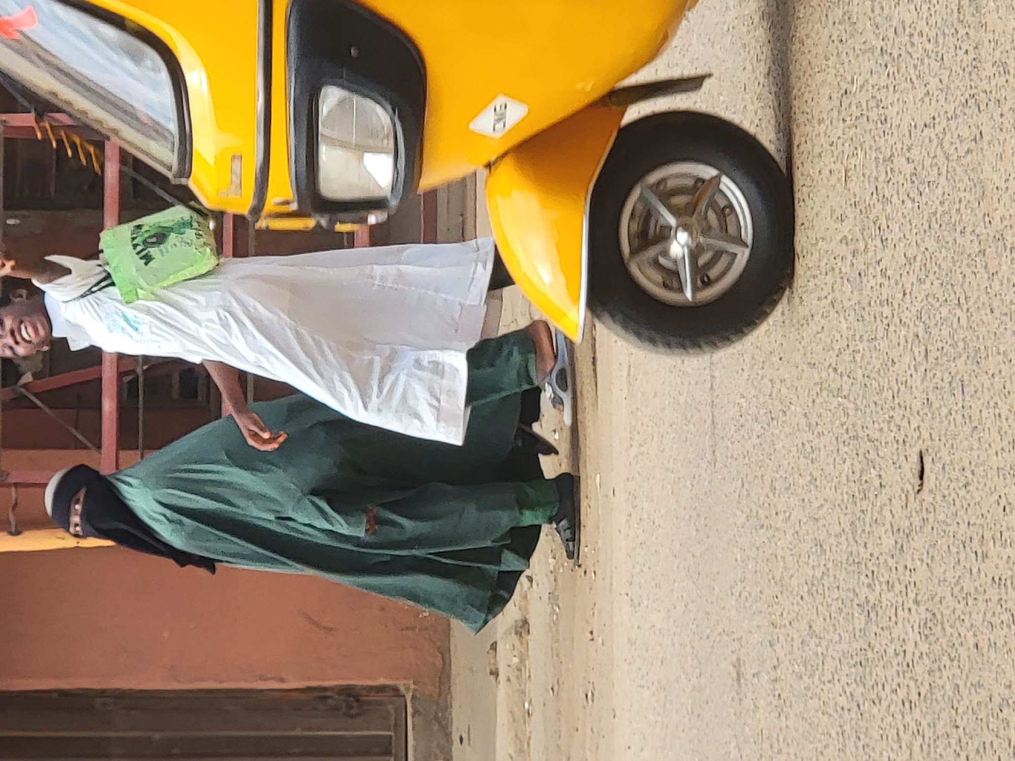 A yellow tricycle is parked on a paved road in Maiburiji, Nigeria. Two people are standing near the vehicle: one wearing a long white garment and carrying a green shopping bag, and the other wearing a dark green garment and a black niqab. A building with a red facade is visible in the background. The road is cracked and shows signs of wear.