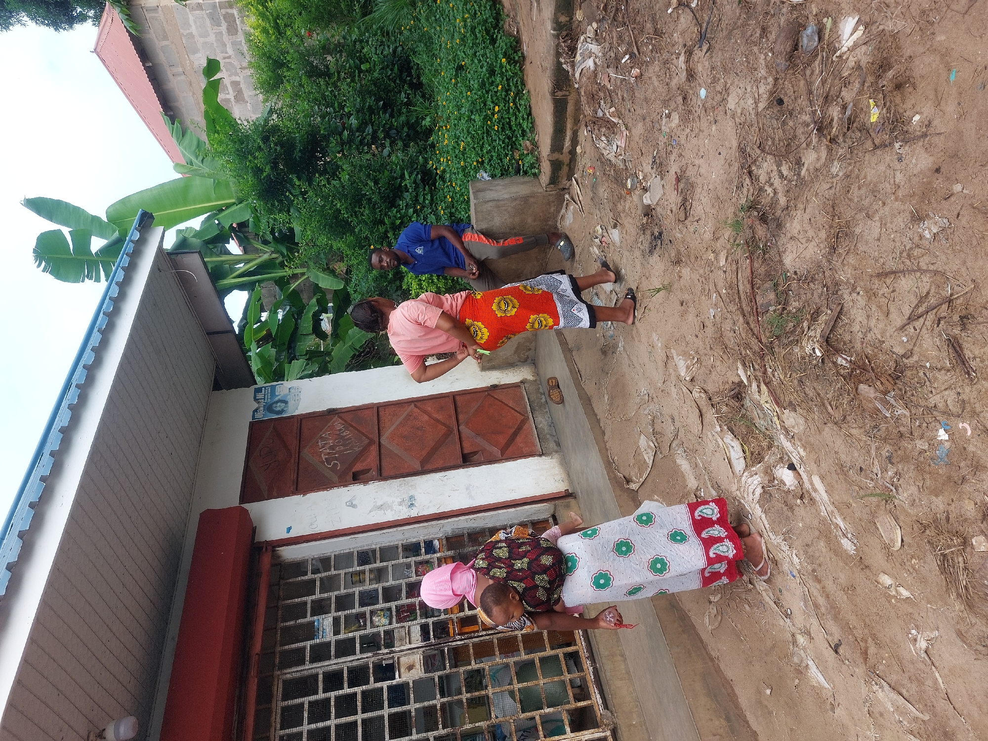 In Dar es-Salaam, Tanzania, the image depicts three people outside a building with a barred window and a red awning. An older woman in a pink head covering, dark patterned top, and white skirt with green flowers stands by the window. A second woman wearing a pink shirt and orange and yellow skirt stands nearby, her hands crossed. A man in a blue shirt and orange pants is seated on a concrete structure behind them. The ground is dirt-covered and littered with debris. Banana trees and greenery are visible behind the building.