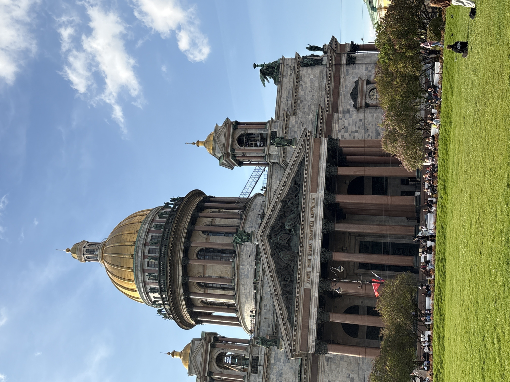The image shows Saint Isaac's Cathedral in Saint Petersburg, Russia. The cathedral is constructed of grey stone with a large golden dome. The image is taken from a low angle and includes a clear blue sky with scattered clouds in the background.
To the right of the cathedral is a green grassy area. Small trees line the periphery, and numerous people sit along a bench bordering the grass. A Russian flag is slightly visible. Some people are walking on the right side of the image. The cathedral is the main subject of the photograph.