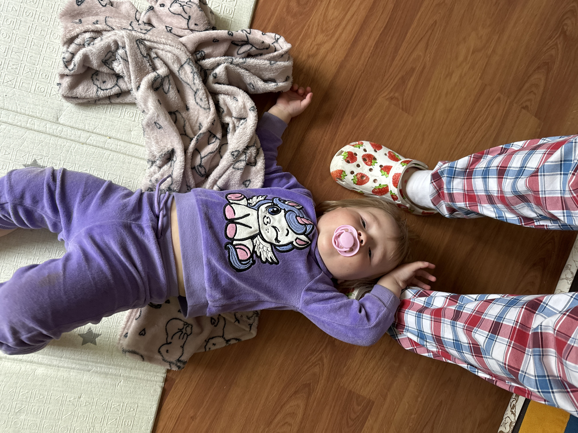 A toddler is lying upside down on a wooden floor, wearing purple sweatpants, a purple sweatshirt with a unicorn design, and a white clog with a strawberry print. The toddler has a pink pacifier in their mouth. Next to the toddler is a pink blanket with animal prints and a light green play mat with letters and numbers. The legs of an adult wearing red, white, and blue plaid pajama pants are visible in the foreground. The scene appears to be indoors, likely in a residential setting. There is no indication of the location being Nizhny Novgorod, Russia.