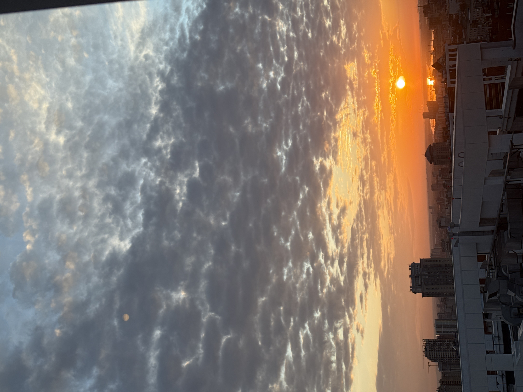 The image depicts a sunrise in Perm, Russia, with a view overlooking a dense urban area. The sky is filled with altocumulus clouds, illuminated by the golden light of the rising sun. The sun is visible as a bright disc near the horizon, casting an orange glow on the sky and buildings below. The city skyline consists of numerous residential and commercial buildings, with some of the structures appearing to be quite tall. The photo seems to have been taken from an elevated position, perhaps from a high-rise building or a hill overlooking the city. There are no visible people or specific actions taking place. The environment is an urban setting with a focus on the atmospheric conditions during sunrise. A portion of the building is at the right side of the image.