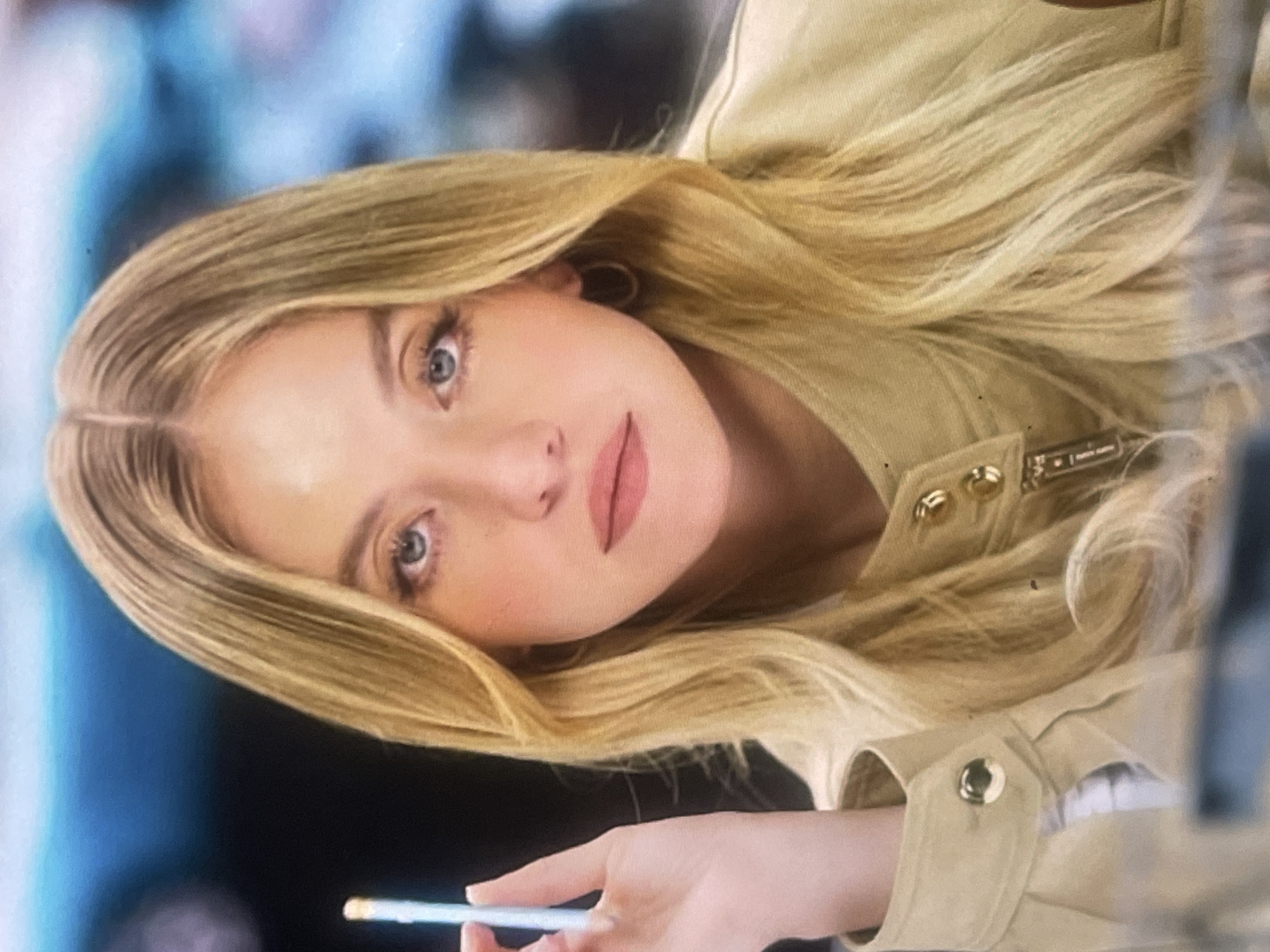 The media file features a young woman with long, straight blonde hair parted in the middle and light blue or grey eyes, looking directly at the camera. Her complexion is fair, and she has visible makeup including blush and a pinkish-nude lipstick. She appears to be wearing a light beige or tan-colored jacket or shirt, possibly made of a sturdy fabric like cotton or linen, with gold-toned buttons on the front placket and cuff. A small, rectangular metallic tag with faint, unreadable text is visible on the jacket near her collarbone, possibly a brand logo. She has a gold hoop earring in her visible left ear.
In her right hand, which is partially visible at the bottom of the frame, she holds a slender, light-colored pen or stylus with a white tip. The background is out of focus, showing blurred areas of blue and darker tones, suggesting an indoor setting or an environment with artificial lighting. The overall impression is that she is engaged in an activity involving writing or digital interaction. Her expression is neutral, with a slight hint of a smile, and a direct, composed gaze. The image's orientation is rotated counter-clockwise. This scene takes place in Poltava, Ukraine.