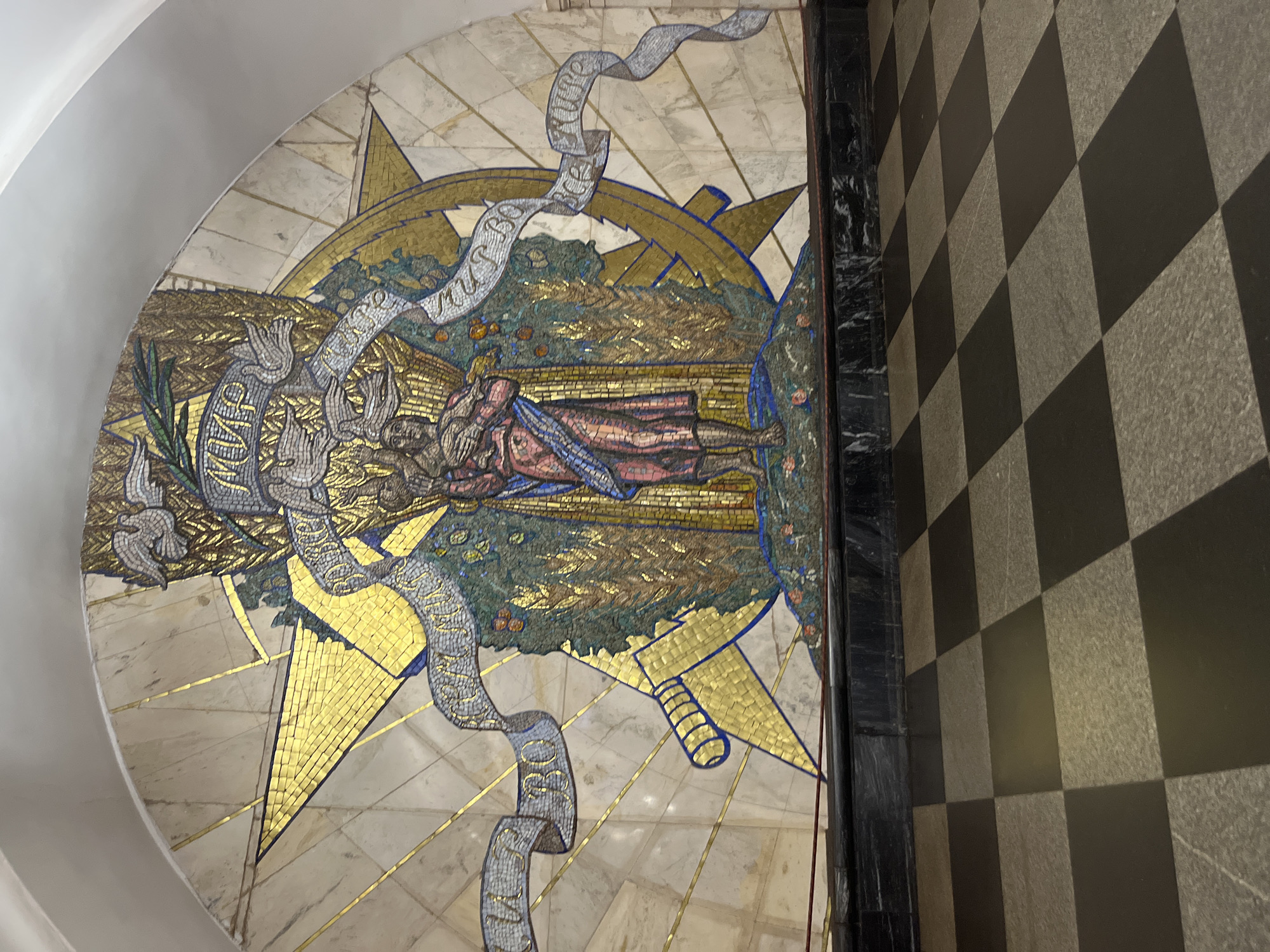 The image shows a large mosaic artwork on a wall, likely in a public space such as a metro station. The mosaic is semicircular and features a central figure holding wheat stalks and a child, surrounded by symbols of peace and prosperity. There are doves, ribbons with text, and a large star. To the left, there appears to be a golden depiction of a bayonet or sword. The background is made of light-colored marble with gold accents radiating outwards from the center. Below the mosaic is a dark stone base, and in the foreground, a black and white checkered tiled floor is visible. This mosaic is located in the Elektrozavodskaya metro station in Moscow, Russia.