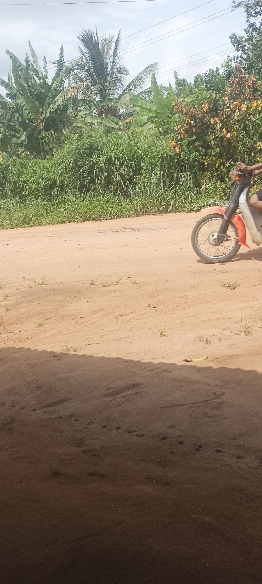 The image depicts a dirt road in Amauzu, Nigeria. Vegetation including banana trees and tall grasses line the roadside. A motorcycle, partially visible, is in motion on the road. The foreground is shaded, possibly indicating the view is from under a structure. The road appears to be sandy or dusty. Power lines are overhead.