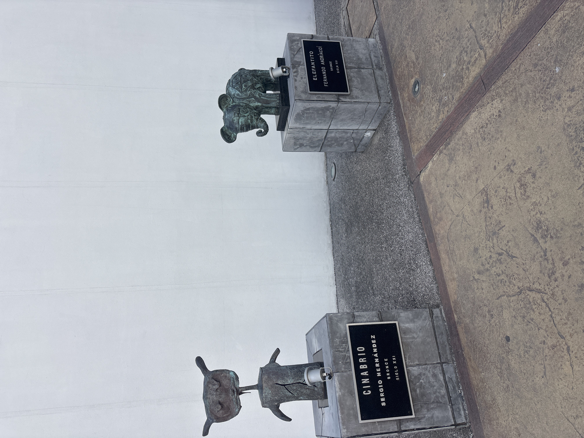 Two bronze sculptures are displayed on concrete pedestals in an outdoor public space within the historic center of Puebla, Mexico, part of the city's "Paseo de Gigantes" art walk.
The sculpture on the left is titled "Cinabrio" by Sergio Hernández, dated 21st Century and made of bronze. It depicts an abstract, humanoid-like figure with a weathered dark patina. The figure has a wide, rounded head featuring two prominent ear-like protrusions and two sunken eye-like depressions. Its body is cylindrical with short, arm-like stumps extending outwards and a visible vertical fissure down the front. This sculpture rests on a dark gray concrete pedestal composed of interlocking blocks. A small, silver-colored spotlight is affixed to the front of the pedestal, pointing upwards.
The sculpture on the right is titled "Elefantito" by Fernando Andriacci, also dated 21st Century and made of bronze. This piece is a stylized representation of an elephant with a dark, weathered patina, covered in geometric patterns. The elephant is depicted standing, facing left, with large round ears and a curved trunk. It is placed on a similar dark gray concrete pedestal, also equipped with a small, silver-colored spotlight pointed upwards.
The background consists of a plain, light-colored (likely white) textured wall with faint vertical seams. The ground in the foreground is a paved surface, segmented into distinct areas. Immediately beneath and surrounding the pedestals, the paving is dark gray and has a coarse aggregate texture. Further into the foreground, the paving transitions to a mottled brown and tan color with irregular patterns, indicative of stamped concrete or large pavers. A reddish-brown strip of paving separates these two primary ground textures. Two small, circular in-ground lights are visible in the brown paved area. No people or overt actions are present in the scene.