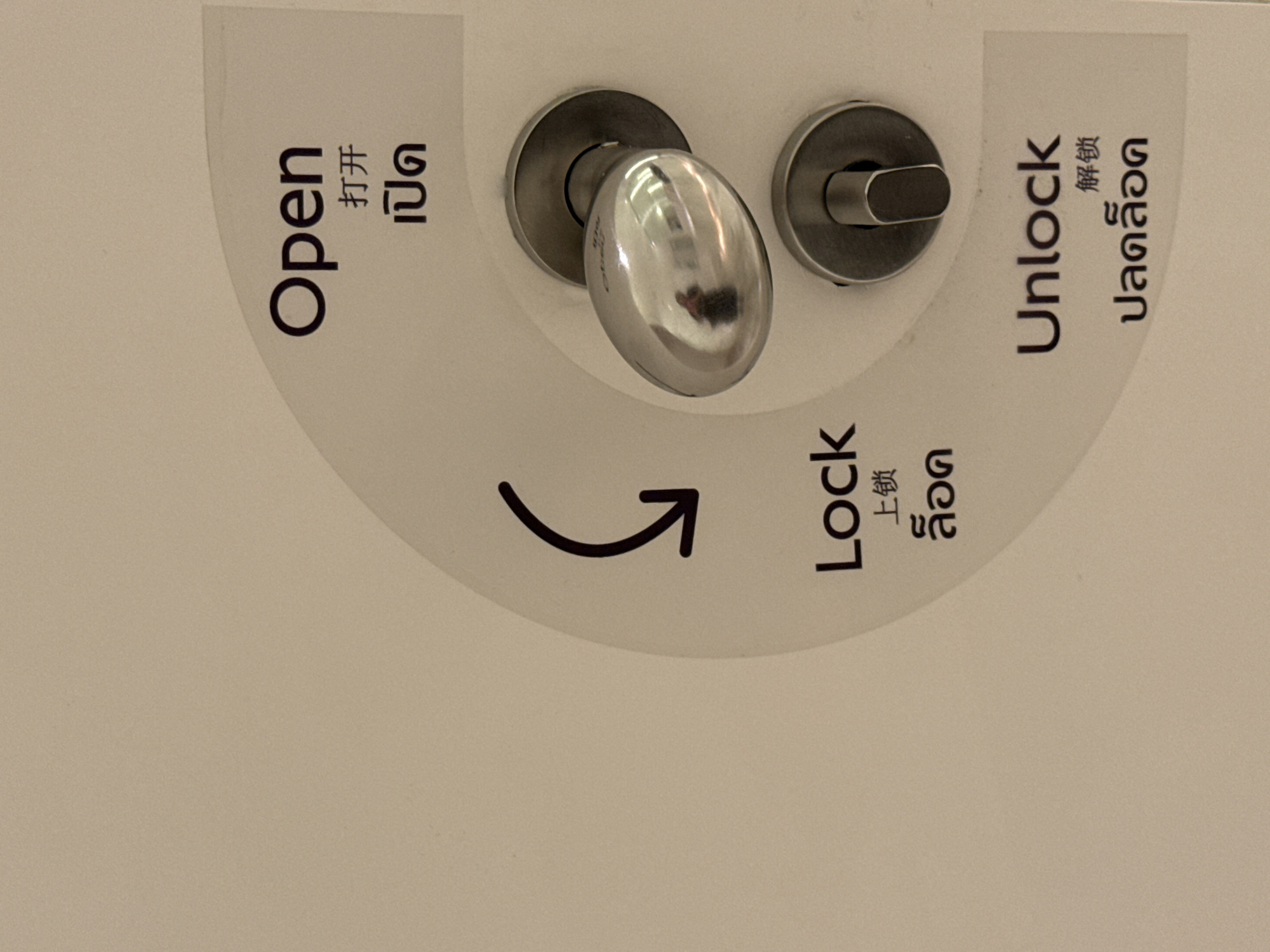 A close-up, slightly rotated image features a modern door handle and an adjacent locking mechanism on a light-colored surface. The central element is a shiny, silver, oval-shaped door knob, reflecting a faint human figure, likely the person taking the photograph. To the right of the handle is a smaller, circular metal piece with a flat, oblong lever used for locking and unlocking.
Around these hardware components, clear operational instructions are provided in English, Chinese, and Thai, printed on a curved, grey-shaded area. The word "Open" (打开, เปิด) is positioned towards the top-left, "Unlock" (解锁, ปลดล็อก) is at the top-right, and "Lock" (上锁, ล็อก) is at the bottom. A curved arrow points from the "Lock" position towards the "Open" direction, illustrating the rotational movement of the handle. The multilingual text, particularly the Thai script, confirms the given context of Bangkok, Thailand, indicating the instructions are for an international audience. The image serves as a guide for operating the door, showing the respective positions for opening, locking, and unlocking.