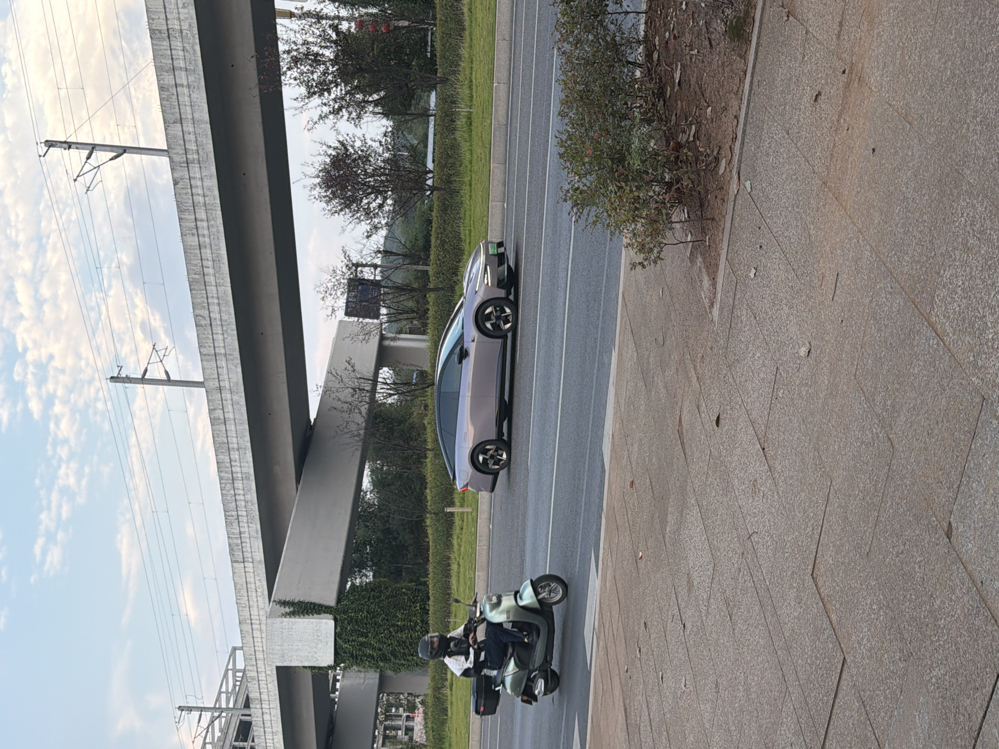 In Jiangjin, China, a light purple electric car travels on a multi-lane road beneath an elevated highway. To the left of the car, a person wearing a helmet and a white and black jacket rides a light green electric scooter, also moving in the same direction. The road surface appears wet, suggesting recent rain or damp conditions. The environment includes green vegetation lining the roadside and a paved pedestrian walkway in the foreground. Overhead, the elevated highway structure, with its concrete supports and overhead power lines for trains, dominates the upper portion of the scene against a sky with scattered clouds.