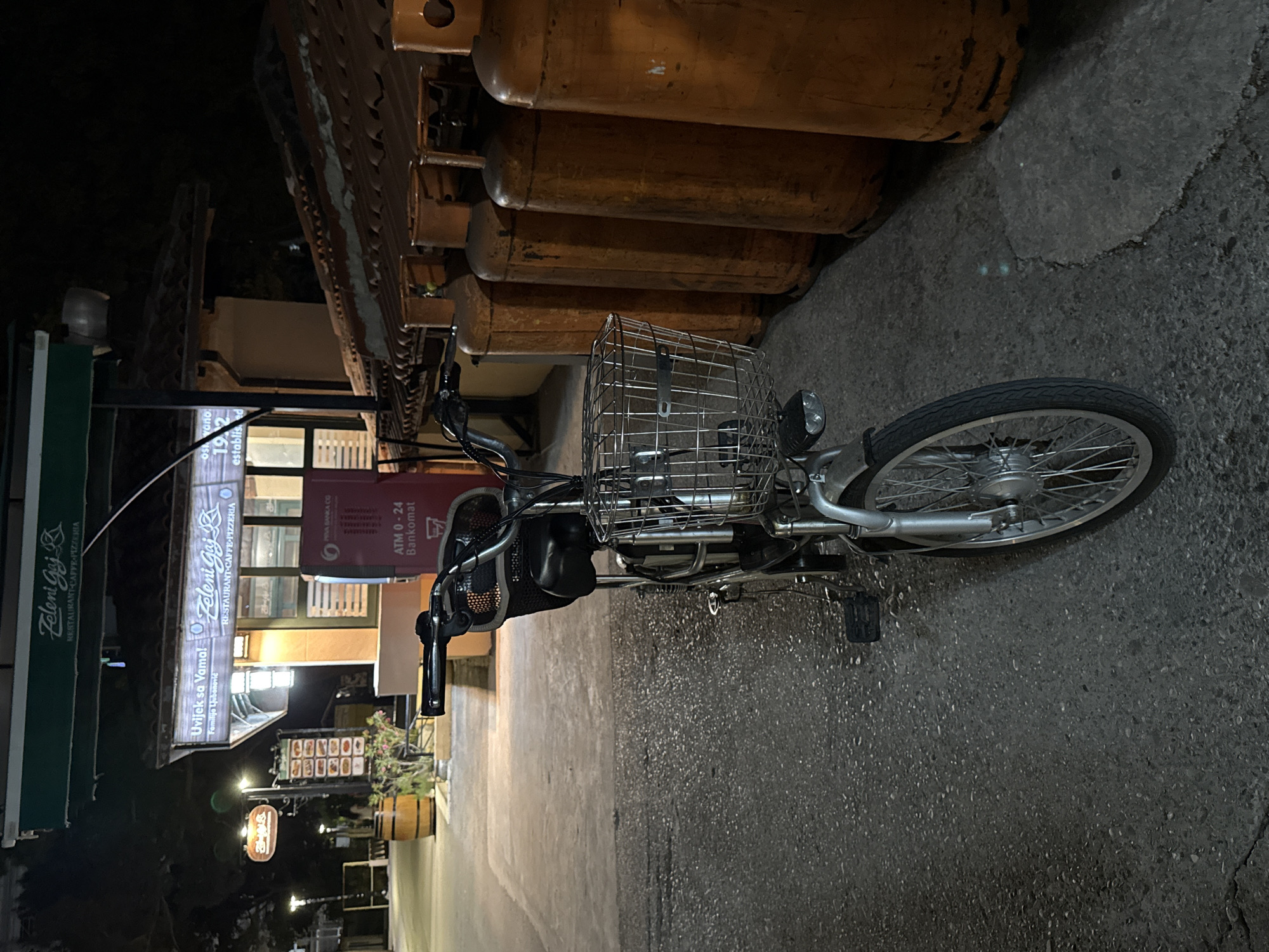 The image captures a scene at night in Budva, Montenegro, outside a establishment identified by signs as "Zeleni Gaj Restaurant-Caffe-Pizzeria." In the foreground, a silver bicycle with a front basket and a rear rack is parked on a paved surface. To the right of the bicycle, a row of large, orange gas cylinders are lined up against a building with a tiled roof. The establishment behind the bicycle has illuminated signage, including one that reads "Uvijek sa Vama! Porodično ljubazno" and another with "Zeleni Gaj RESTAURANT-CAFFE-PIZZERIA." A red ATM machine is visible to the right of the main entrance. The overall environment suggests an outdoor seating area or a service entrance of a commercial establishment at night.