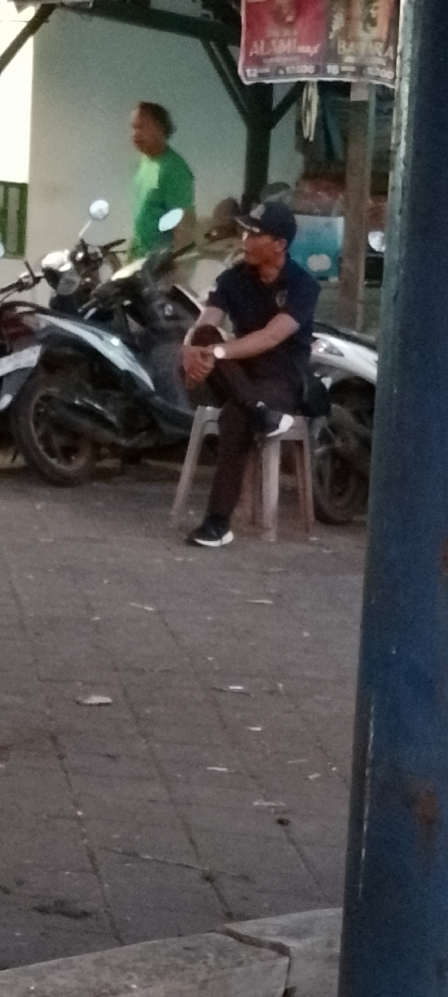 In Pekutatan, Indonesia, a man in a dark uniform and a dark cap is seated on a light-colored plastic stool. He is wearing dark pants and dark shoes with white accents. He has his arms crossed and his legs are crossed at the knees. To his left, there are two parked motorcycles. In the background, another person in a green shirt is visible. A poster is hanging above the motorcycles. The ground is paved with interlocking stones. A large blue pole is on the right side of the frame.