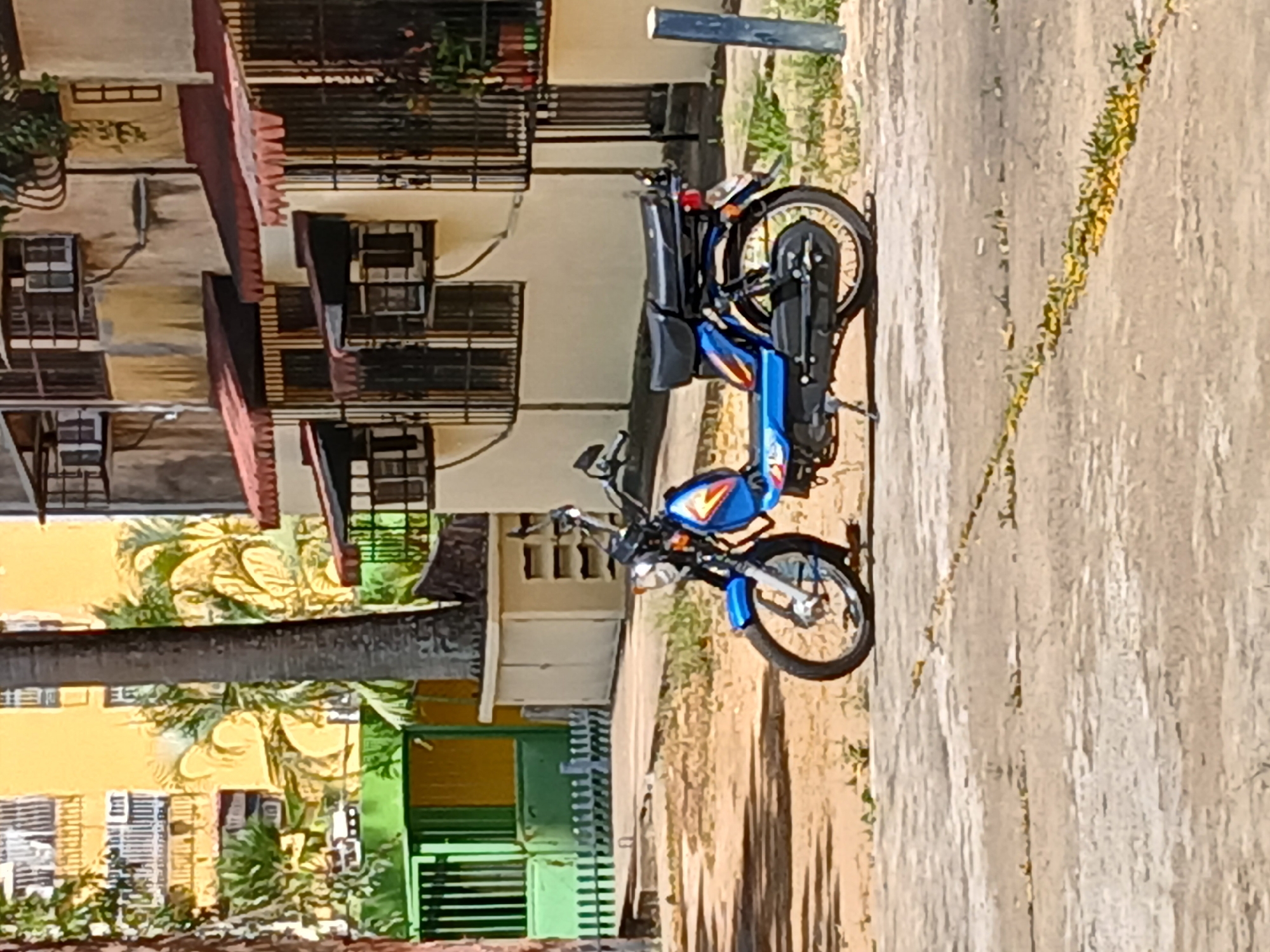 An outdoor, sunlit scene depicts a blue and black moped or small motorcycle parked on a paved surface, likely a pathway or small street, in Ciudad Guayana, Venezuela. The image, originally rotated, shows the vehicle positioned prominently on the right side of the frame, facing towards the left.
The moped, the central subject, features a vibrant blue body with a black seat, handlebars, and frame. It has a small windscreen and a functional cargo rack over its rear wheel. The fuel tank displays colorful decals in shades of red, yellow, and orange, along with a partially visible but illegible logo or text. The vehicle casts a distinct shadow on the light-colored pavement, indicating bright, direct sunlight.
In the background, a series of multi-story buildings with light-colored (beige or yellowish) facades are visible, suggesting a residential or mixed-use neighborhood. These structures feature numerous windows, some with security grates or dark frames, and are accented with reddish-brown tiled roofs or awnings. Lush green foliage, including palm trees, is interspersed among and behind the buildings, with smaller plants growing along the edges and cracks of the paved area. A distinctive green metal fence or gate is also noticeable in the lower-left background, and a dark, slender pole stands vertically near the edge of the paved surface.
The time of day appears to be mid-day or early afternoon, characterized by strong shadows and clear, bright lighting, suggesting sunny weather. No people or specific activities are depicted; the scene is static.