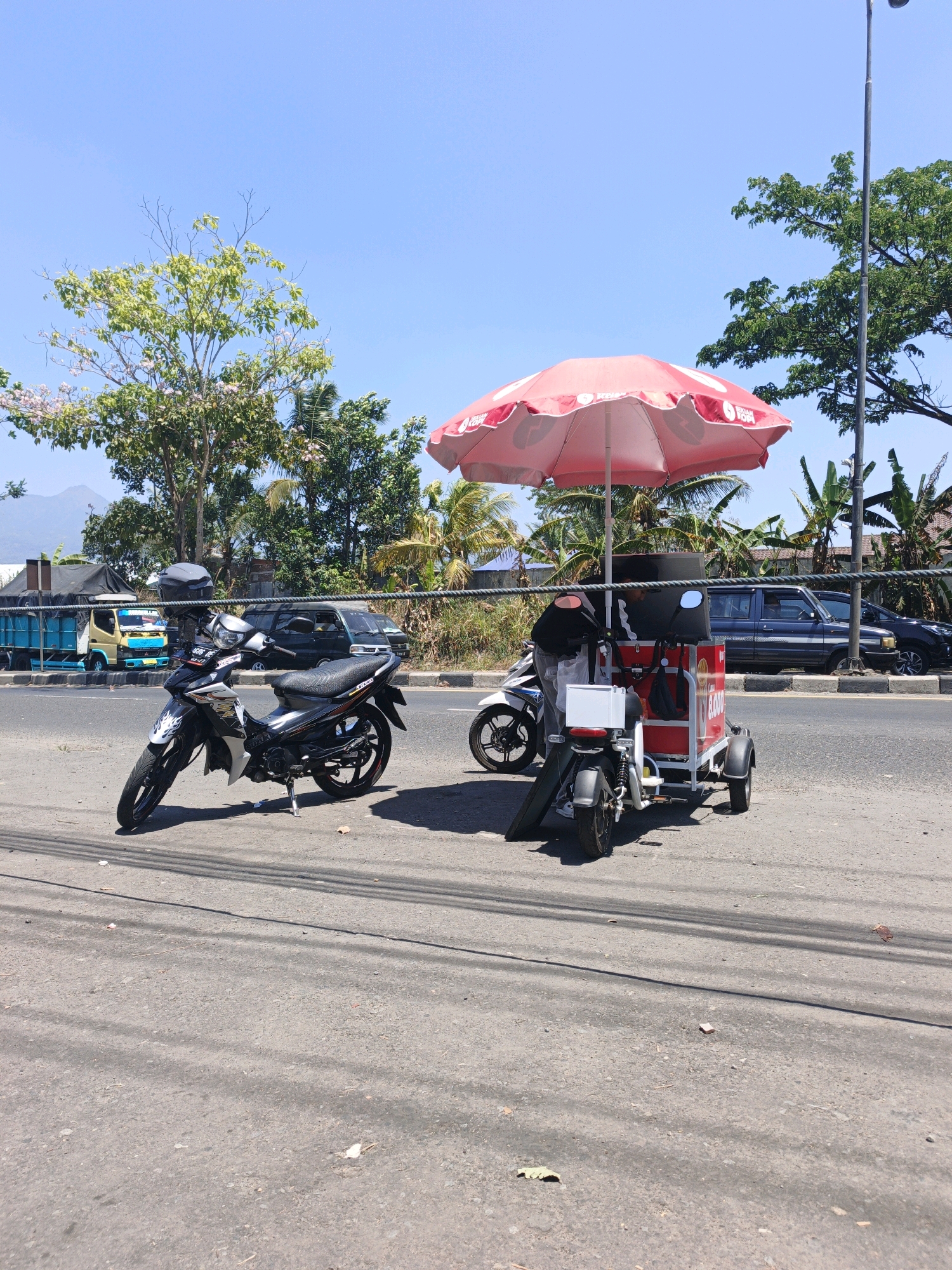 In an outdoor, urban setting under a clear blue sky, two motorcycles and a three-wheeled cart are parked on the side of a paved road. The foreground features a black and white motorcycle, parked upright. To its right, a smaller, white electric motorcycle is parked next to a red and white cart, which is topped with a large, pink umbrella. A person, partially visible, is interacting with the cart. In the background, several vehicles, including a blue truck and a dark SUV, are on the road. Lush green trees and vegetation line the roadside, with a distant mountain visible on the left. The overall scene depicts a roadside scene with vehicles and a mobile vendor.