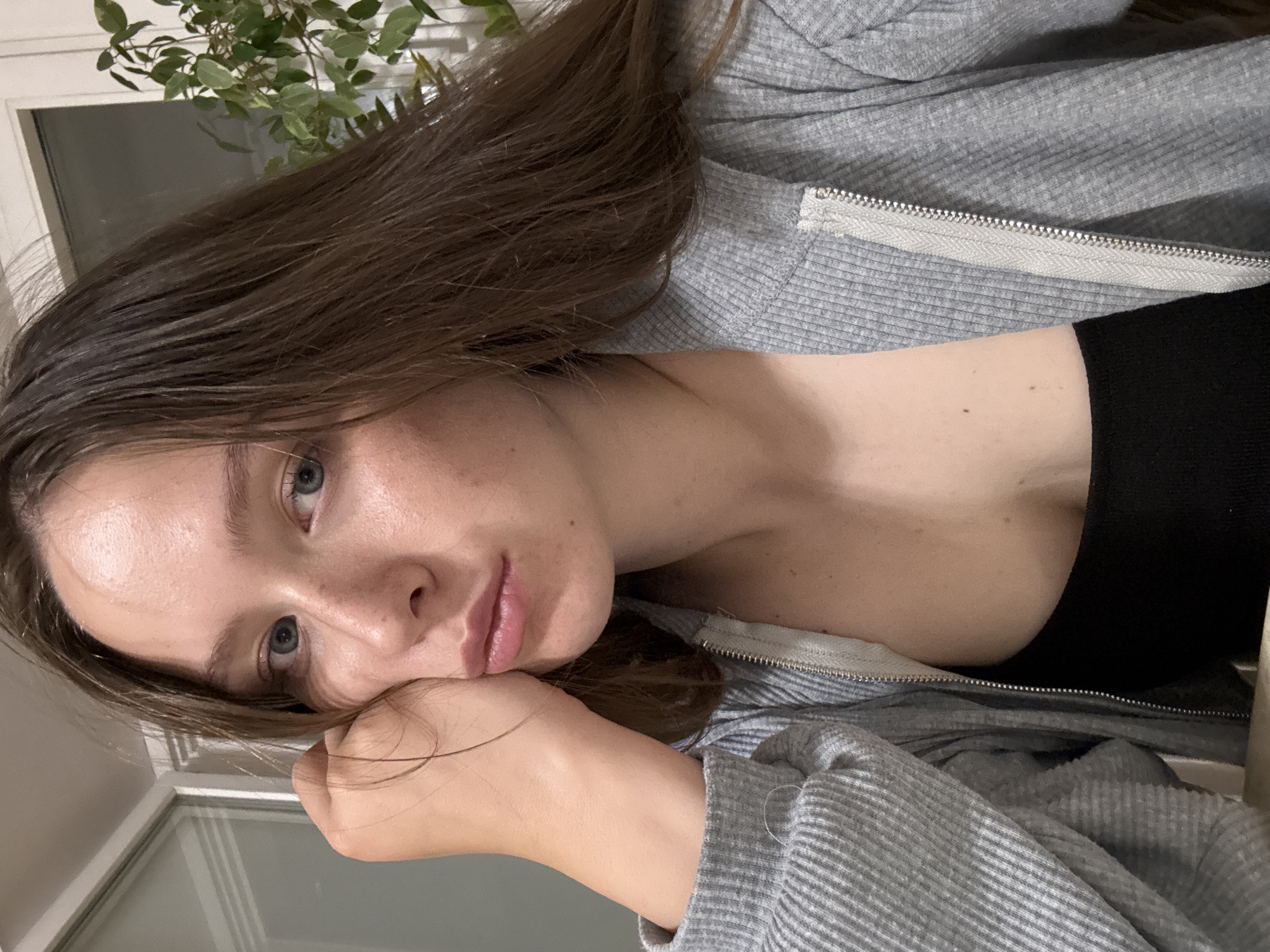 A young woman with light skin, brown hair, and blue eyes is resting her head on her hand, looking at the camera. She is wearing a grey zip-up sweater and a black top. In the background is a window with a plant on the windowsill. There is no evidence in the image to determine the location as Chaikovsky, Russia, or any other specific location.