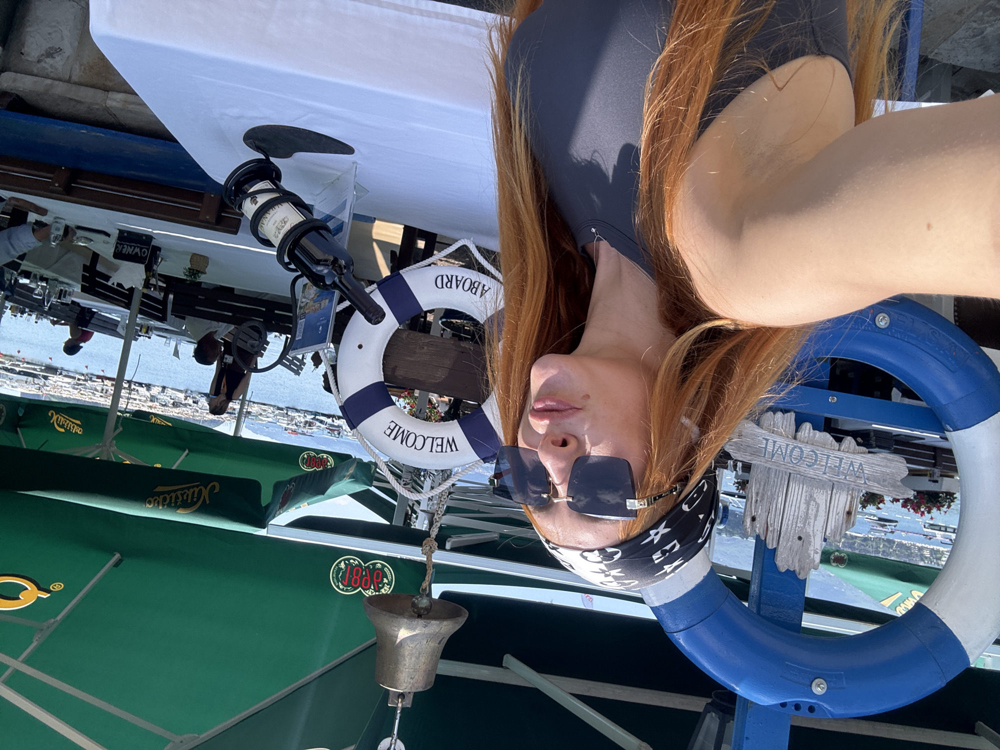 A woman with long, red hair and a bandana is taking a selfie in an outdoor dining area near a harbor. She is wearing sunglasses and a dark gray top. In the foreground, a blue and white life preserver with the word "WELCOME" on it is visible. Behind her, a bell hangs from a chain. Several tables with white tablecloths are set up, and people are seated at some of them. Green umbrellas provide shade over the tables. Boats are docked in the harbor in the background, and mountains are visible in the distance. The scene appears to be in Budva, Montenegro, a coastal town known for its historical sites and beaches.