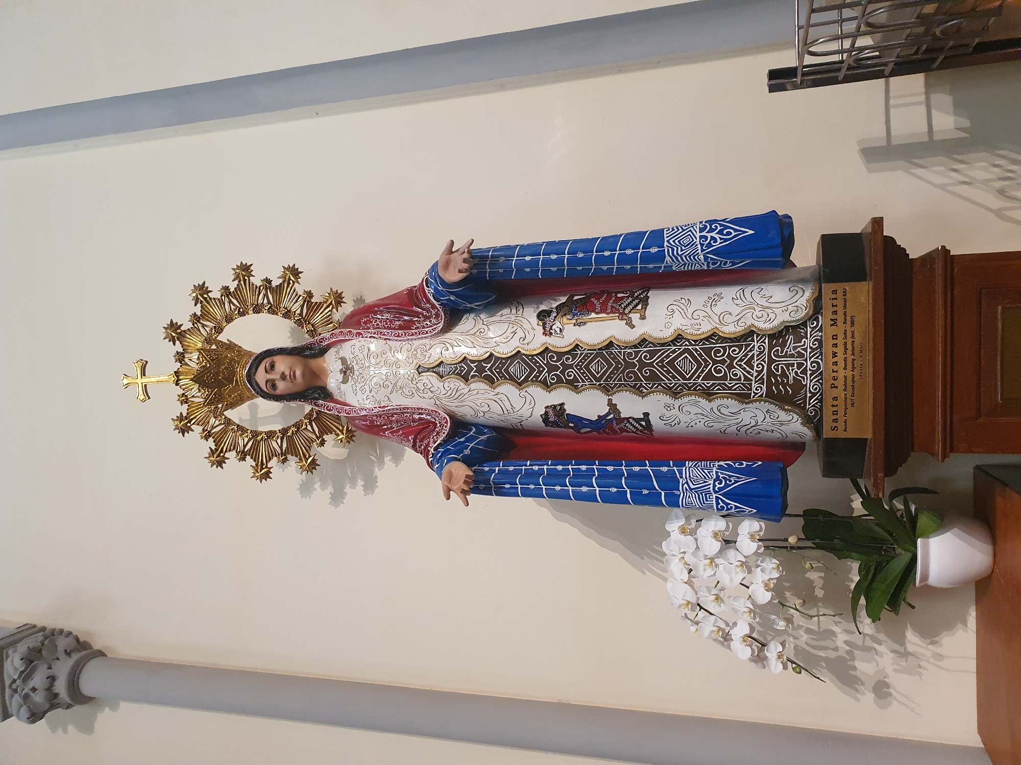 A full-body, eye-level shot depicts a statue of Mary in a church. The statue, standing on a wooden pedestal, is adorned with a golden crown and halo, a white and gold patterned dress, and a red and blue mantle. The mantle is intricately decorated with blue and white checkered patterns and appears to be made of cloth. White orchids in a white pot are placed to the left of the statue. The background consists of a plain, light-colored wall with vertical grey and blue stripes, and a column on the left. A metal grate is visible to the right of the statue. The sign at the base of the statue reads "SANTA PERAWAN MARIA," "Rahmat - Bunda Segala Suku - Bunda Umat KAJ," "Keuskupan Agung Jakarta (est. 1807)," and "(Pesta 8 Mei)." This suggests the statue is located in the Archdiocese of Jakarta, Indonesia, and commemorates May 8th as a feast day.