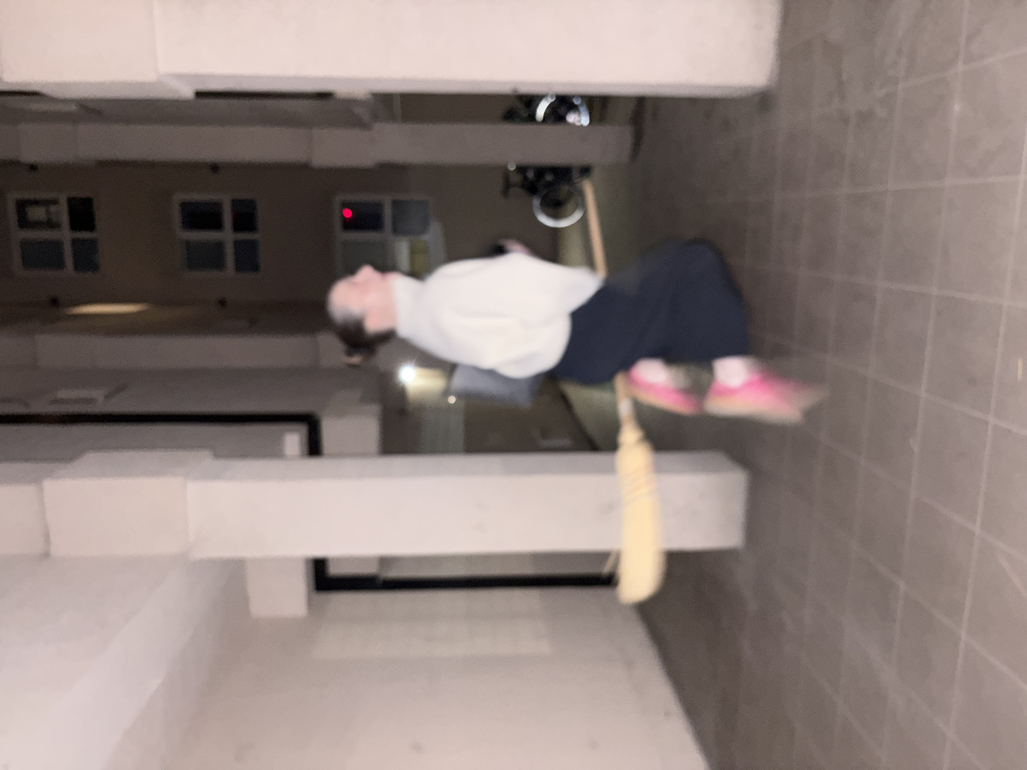 A low-quality, blurry photograph depicts a person, likely female, lying prone on what appears to be a tiled surface. She is wearing a light-colored top, dark pants, and pink shoes. A broom is positioned near her, handle extending toward her. The environment features architectural elements suggestive of a building's exterior. Windows are visible in the background. Based on these details, and the request for the location, this scene is set in Kragujevac, Serbia. However, the specific location is impossible to identify without further context.