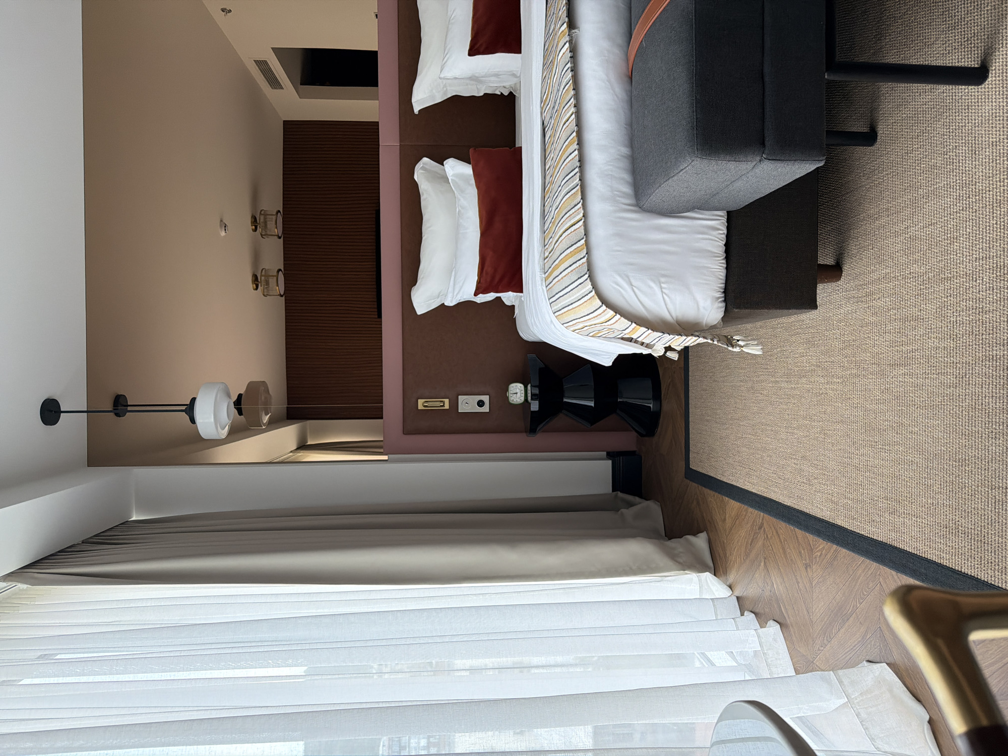 This is a medium shot, eye-level, interior photograph of a hotel bedroom. The room features a large bed with white linens, a dark red bolster pillow, and white pillows. A striped throw is draped across the foot of the bed. A dark grey upholstered bench with leather straps sits in front of the bed. To the left of the bed, sheer white curtains are drawn back, revealing a glimpse of a city view. A dark, sculptural side table with a small green alarm clock sits next to the bed. The room has wood-paneled walls behind the headboard and a modern pendant light fixture hangs from the ceiling. The flooring is a herringbone wood pattern covered in part by a natural fiber rug with a dark border. The overall impression is one of modern luxury. This hotel room is likely located in the city of Tbilisi, Georgia.