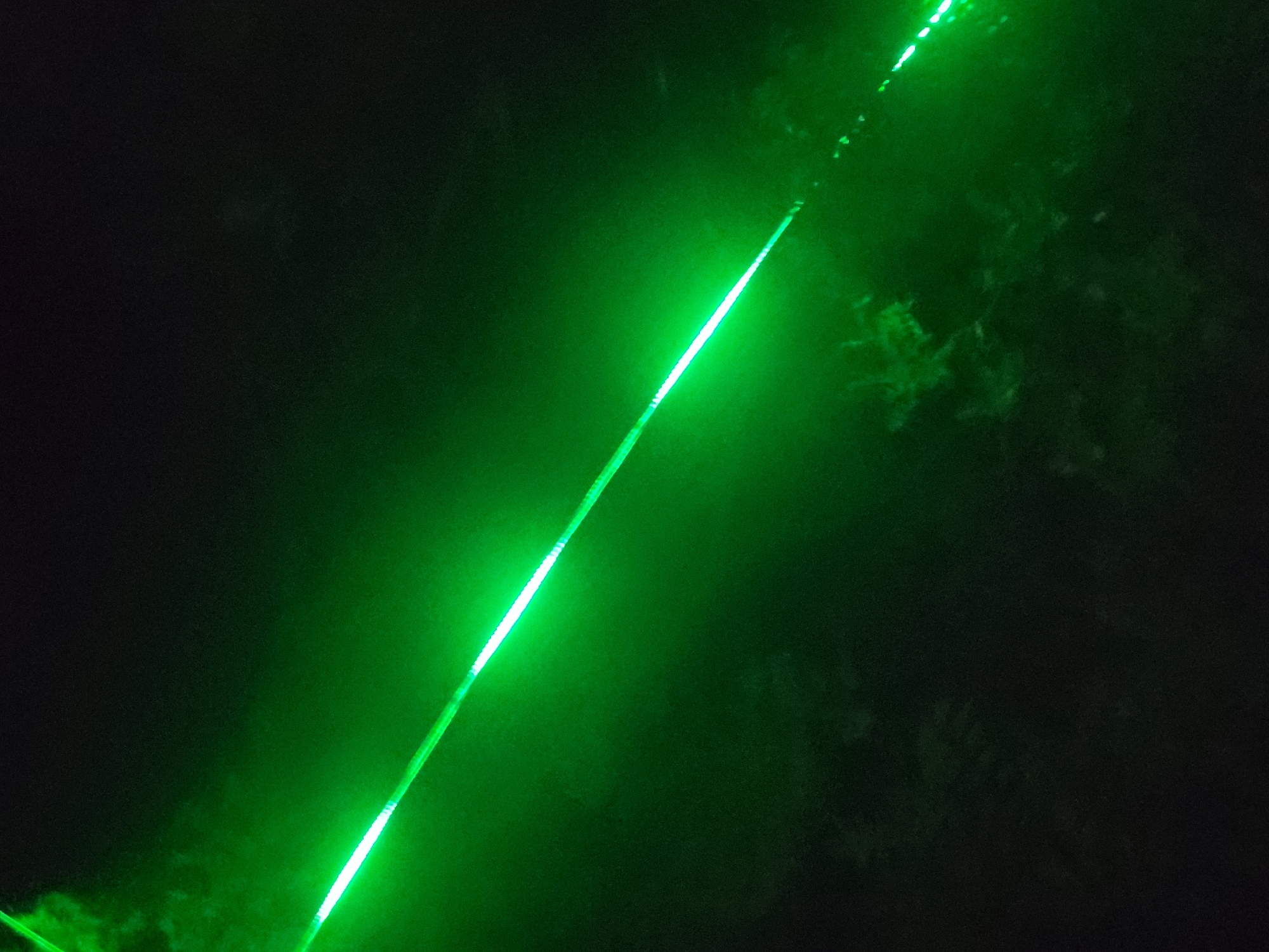 A dark, low-light photograph captures a scene at night in Keffi, Nigeria. The central feature is a prominent, bright green diagonal line composed of distinct, illuminated segments. This string of lights originates from the upper-left frame edge and extends towards the lower-right frame edge, running across the majority of the image. The individual light segments are short, bright green, and appear to be arranged in a dashed pattern along a continuous medium, suggesting a string of LEDs or similar light fixture rather than a continuous beam.
The surrounding environment is characterized by near-total darkness, indicative of a nocturnal setting. Faint, indistinct dark forms, consistent with tree foliage or bushes, are discernible in the background, particularly in the upper-left and central-right sections, subtly illuminated by the ambient green light spill. The image exhibits significant visual noise and grain, typical of low-light photographic conditions. No individuals, specific man-made objects beyond the light string, or discernible human actions or interactions are present within the frame.