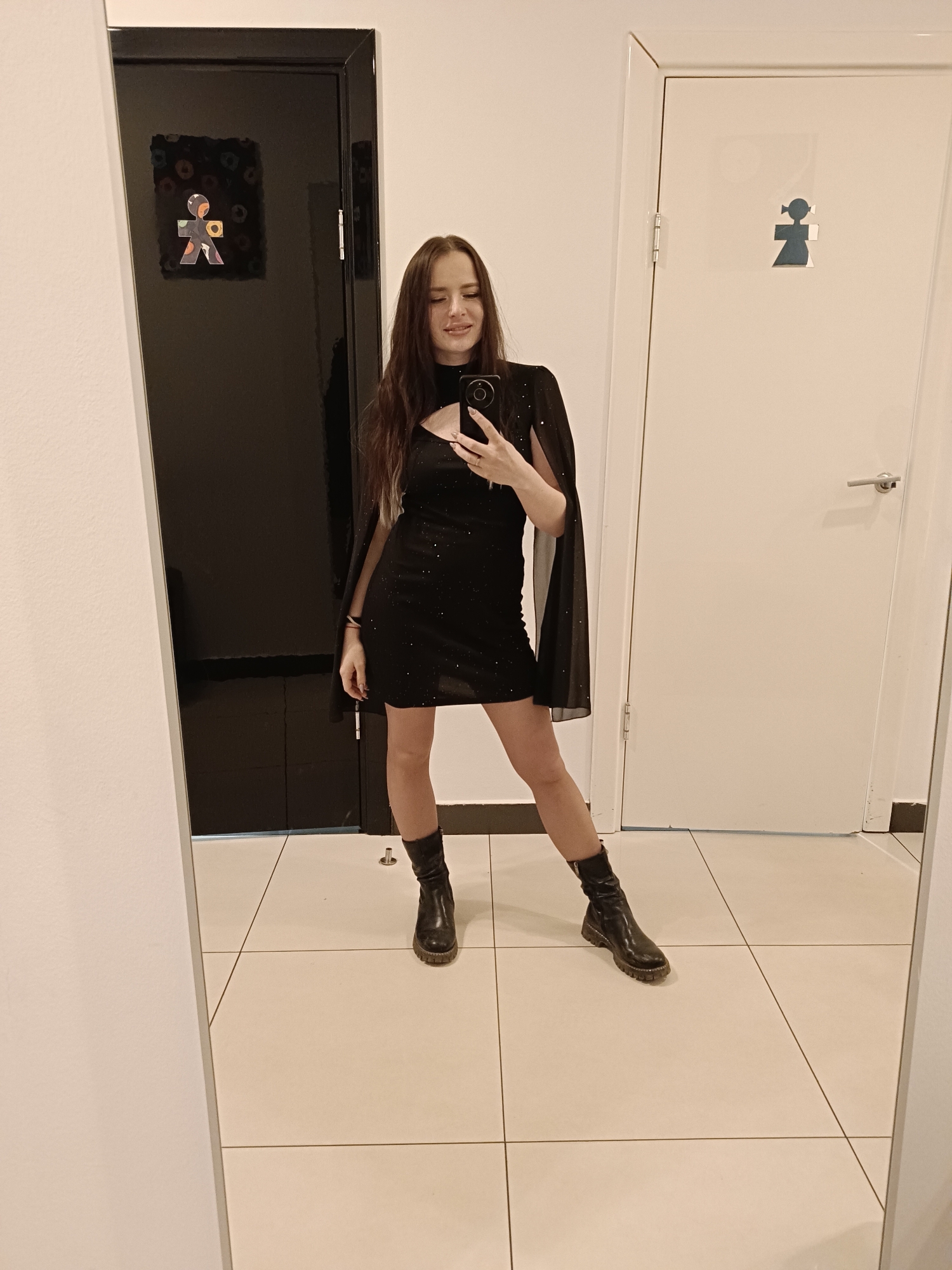 A woman is standing in front of a mirror, taking a selfie. She is wearing a black mini dress with a cape and black boots. The location appears to be a restroom or dressing room with two doors. One door is black with a stylized human figure; the other is white with a more traditional female restroom symbol. The floor is tiled. The exact location within Chaikovsky, Russia, cannot be determined from the image.