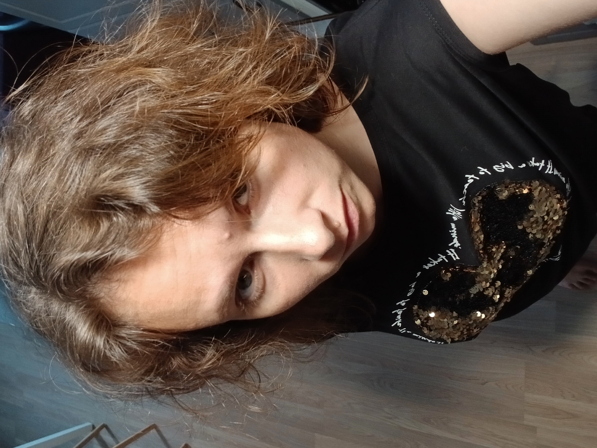 The image shows a woman from the chest up, looking directly at the camera. She has medium-length, wavy brown hair. Her face is pale and her eyes are open. She is wearing a black t-shirt with a heart-shaped design made of gold sequins on the front, surrounded by white text. The background appears to be an indoor setting with wooden flooring and a partially visible furniture item. There are no indications of the specific location in Yekaterinburg, Russia.
