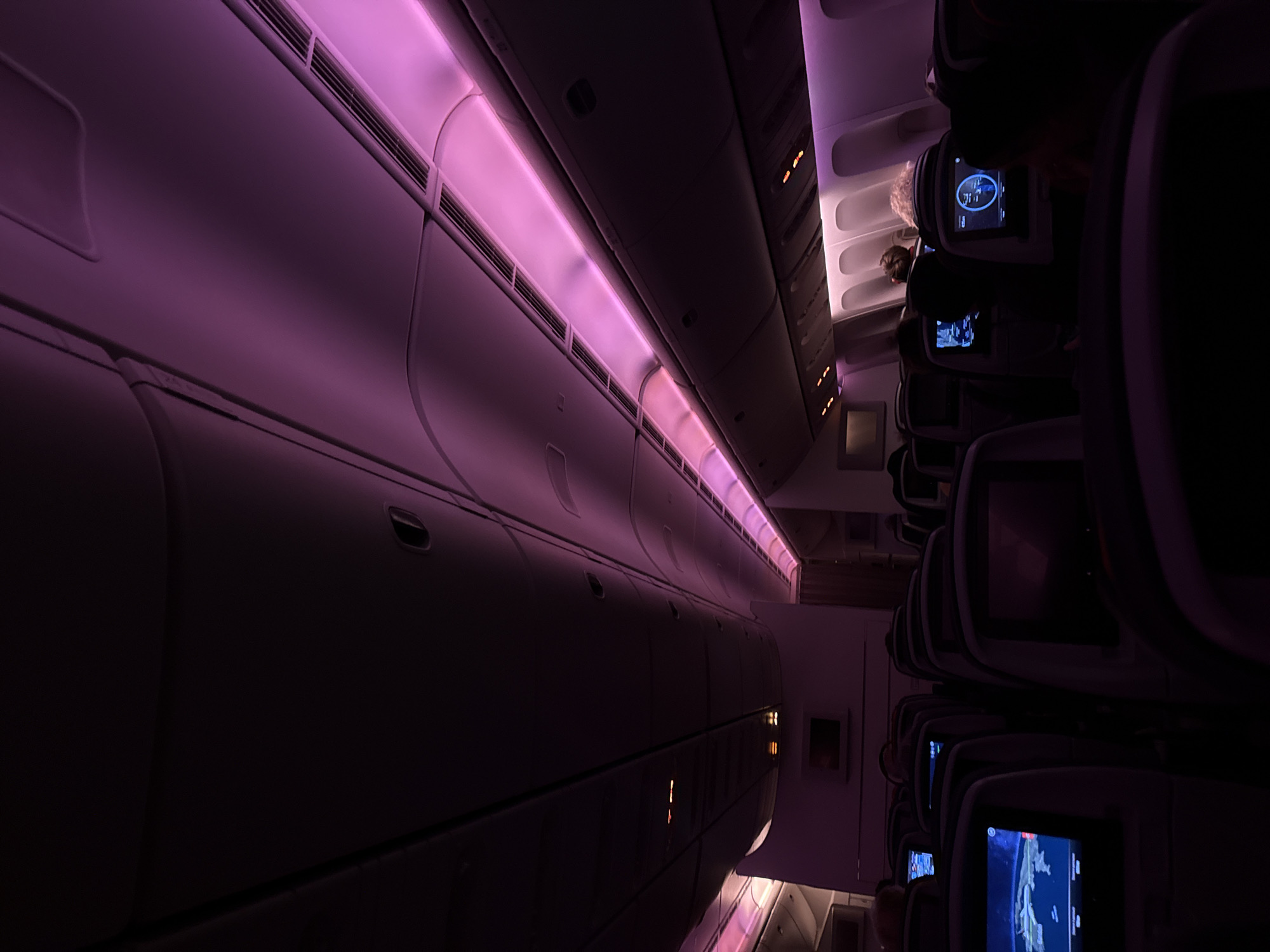 The image shows the interior of an airplane cabin. Rows of seats are visible on the right, with passenger entertainment screens on the back of each seat. The interior is lit with purple lighting along the ceiling and storage compartments on the left. Passengers can be seen in some of the seats. The location is not visible in this image, so determining if it is in Clichy, France is impossible.