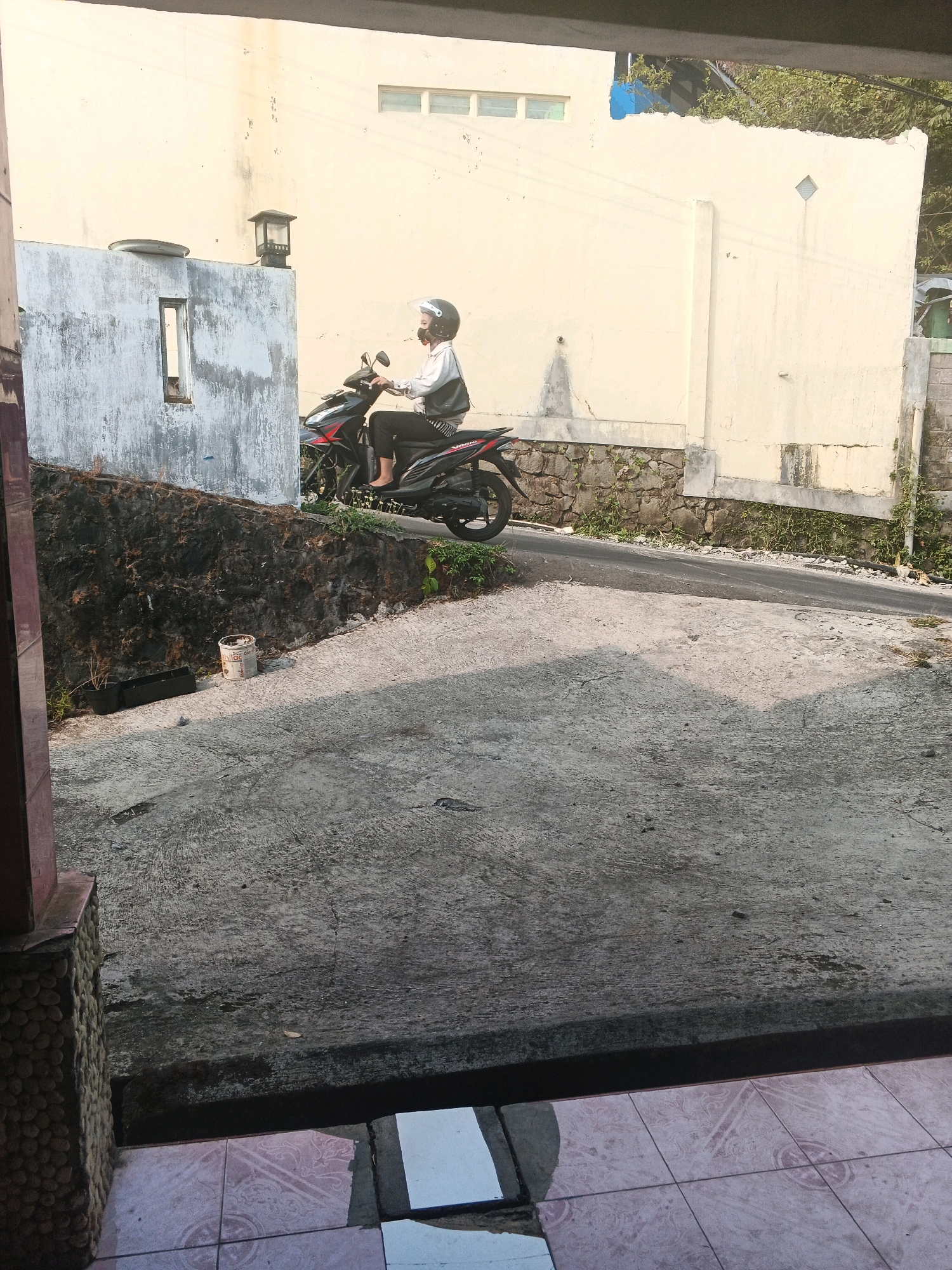 A side profile, full shot, captures a person wearing a helmet and a white shirt over dark clothing, riding a black and red motorcycle on an inclined asphalt road. The person is positioned on the right side of the frame. Behind the motorcycle, a textured stone wall with patches of greenery runs horizontally, supporting a light yellow building with horizontal window slats. To the left of the motorcycle, a grey concrete structure with a rectangular opening is visible. In the foreground, a textured concrete surface slopes downwards, with some debris and a small, dark pot on the left. The scene appears to be set outdoors, with natural light casting shadows, suggesting a daytime setting in Keji, Indonesia.