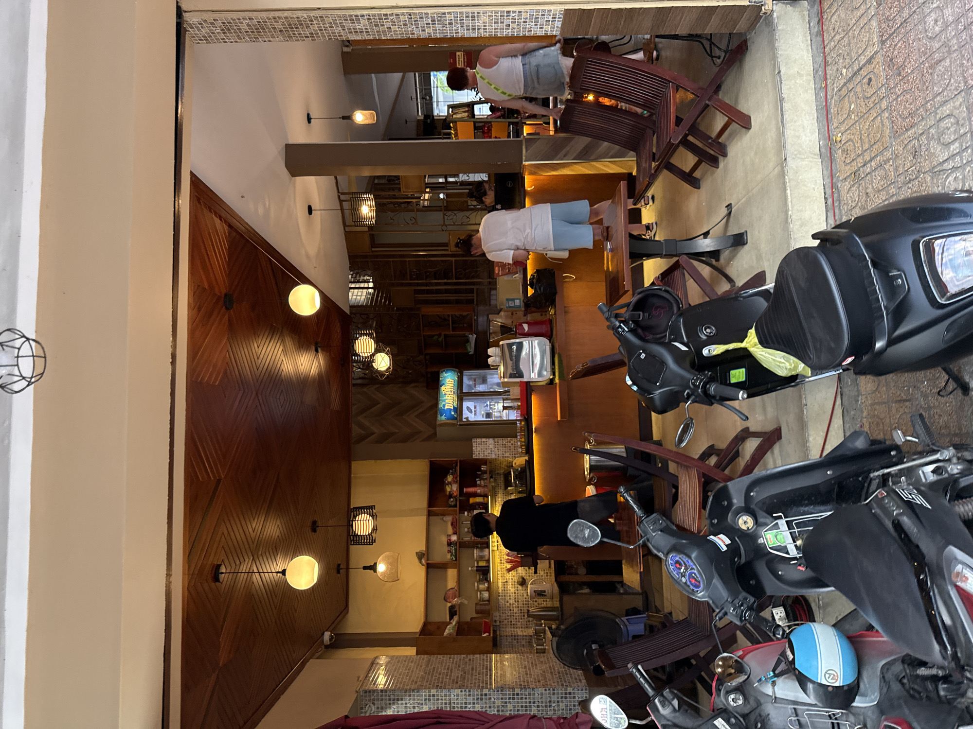 The image shows the exterior of a street-level cafe in Nha Trang, Vietnam, with motorbikes parked along the sidewalk in the foreground. The cafe has an open-air seating area and an indoor counter area. Inside, there are people working and customers present. The cafe has a wooden ceiling and a wooden counter. A refrigerator displaying drinks is visible behind the counter. Various cafe equipment, such as a coffee machine, are also present. Several wooden chairs and tables are set up for customers. The floor is tiled. The scene appears to be during daylight hours.