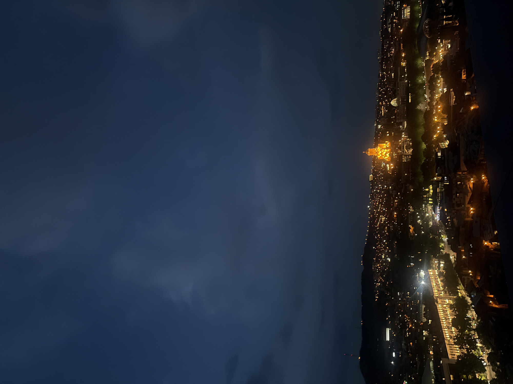 The image captures an elevated view of Tbilisi, Georgia at night. The sky is dark, transitioning from a lighter blue-gray near the horizon to a deeper blue-black above. City lights illuminate the landscape, with a prominent, brightly lit structure resembling a cathedral or large church standing out. Smaller, scattered lights mark buildings and streets, creating a network of brightness across the cityscape. The overall scene conveys a sense of urban activity in the evening. The Holy Trinity Cathedral of Tbilisi, or Sameba, is highly likely the church in view given the size and illumination, confirming the location as Tbilisi, Georgia.