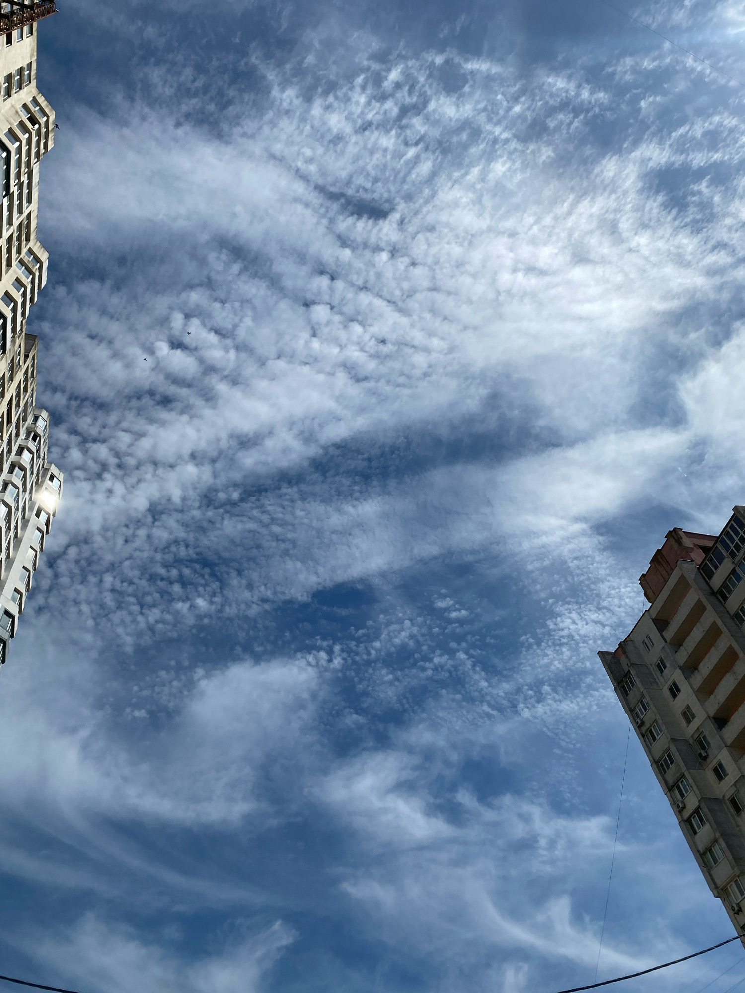 The image shows two apartment buildings in Chornomorsk, Ukraine, against a blue sky filled with scattered white clouds. The building on the left is lighter in color and appears to have more windows. The building on the right is darker and shows balconies. The perspective is looking upwards from street level, with power lines visible at the bottom of the frame. There are no people or animals visible in the image.