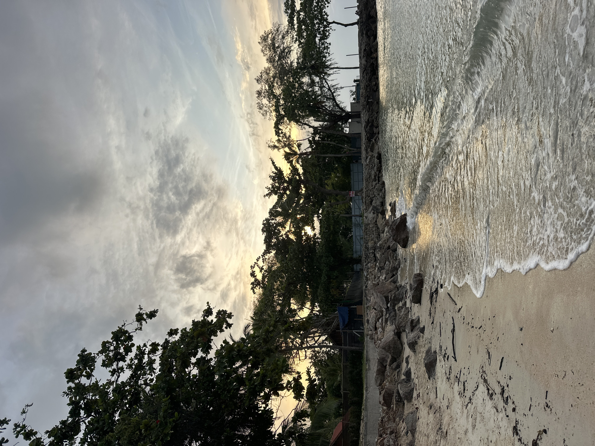 The image depicts a tranquil beach scene in Ko Samui District, Thailand, at dusk. The foreground is dominated by a sandy beach littered with small stones and driftwood, with a gentle wave lapping at the shore. The water reflects the warm hues of the setting sun, creating a shimmering effect. Beyond the beach, a rocky embankment separates the sand from a line of lush green trees and palm trees. In the background, several structures are visible, including a small building with a red roof, a blue tarp-covered shelter, and fencing. The sky is a canvas of dramatic clouds, with streaks of orange and yellow indicating the sunset. The overall atmosphere is one of peaceful serenity.