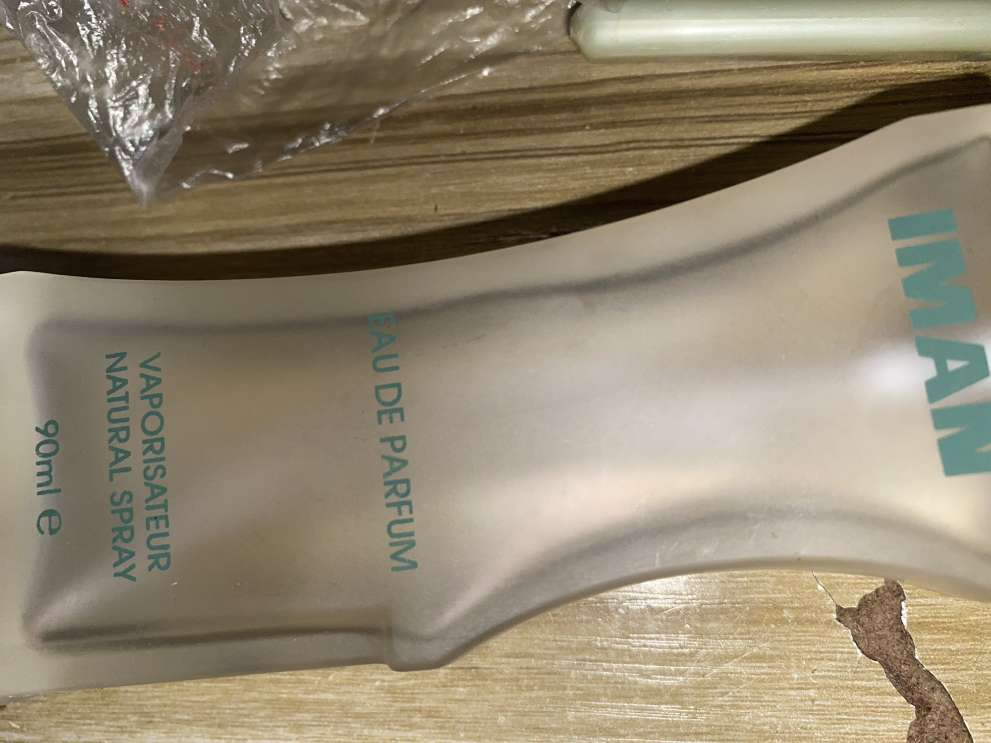 An object, possibly a bottle of perfume, is shown in the image. The object is a light-colored, translucent material with text printed on it in a teal color. The text reads "90ml e", "VAPORISATEUR NATURAL SPRAY", and "EAU DE PARFUM". Below this, the text "IMAN" is visible. The object is set against a background of a wood-grain surface. To the right of the object, a portion of a transparent plastic wrapper with red material visible inside is present. Further to the right, a cylindrical object, light in color, is partially visible. The image appears to be a close-up shot of these items.