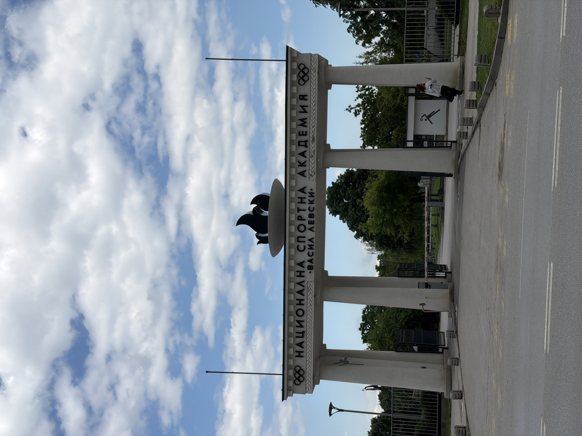 The image displays the grand entrance gate of the National Sports Academy "Vasil Levski" in Sofia, Bulgaria, under a bright, partly cloudy sky. A large, light-colored archway serves as the main entrance, supported by multiple pillars. Along the top beam of the archway, the name "НАЦИОНАЛНА СПОРТНА АКАДЕМИЯ "ВАСИЛ ЛЕВСКИ"" is written in Cyrillic script, meaning "National Sports Academy "Vasil Levski"". The Olympic Rings symbol is prominently featured on both ends of the textual sign, and a stylized black sculpture, resembling a bird or flame, tops the central part of the arch.
To the right of the entrance gate, a single person is visible, dressed in a light-colored jacket and dark pants, holding what appears to be a phone up towards their face. Behind them, a white rectangular sign displays a black pictogram of a figure involved in a sport, possibly with a ball. The foreground features an asphalt road with clear white and yellow markings, leading up to the academy. Small concrete bollards line the edge of the road near the gate. In the background and beyond the gate, lush green trees are visible, along with metal fences and a red-and-white barrier arm, indicating a controlled entry point. The scene is illuminated by natural daylight, suggesting it is either late morning or afternoon. Two flagpoles stand tall on either side of the archway, though no flags are present.