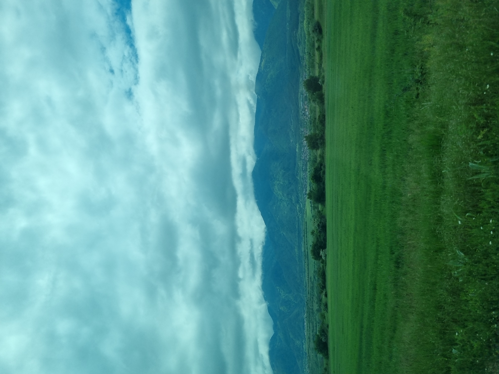 The image shows a landscape with a bright, cloudy sky covering approximately half the frame. Below the sky, there is a row of mountains that stretch horizontally across the middle of the image. Adjacent to the mountains, there is a strip of green land, appearing as a field or meadow. The field extends to the right edge of the frame. There is no evidence of Yahinovo, Bulgaria. There are no people or interactions visible in the image.