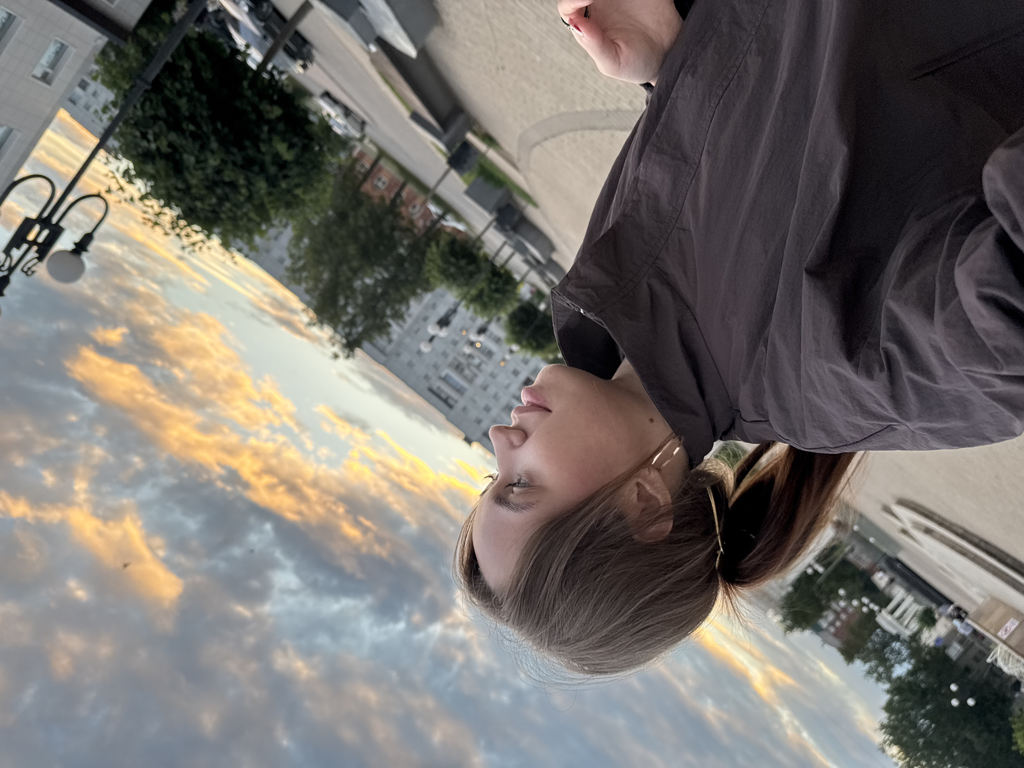 The image shows a young woman with brown hair pulled into a ponytail, wearing large hoop earrings and a dark jacket. She is looking upwards towards a sky filled with dramatic clouds illuminated by a sunset. The background depicts a cityscape with buildings, trees, a street with cars, and a lamppost, consistent with an urban setting in Chaikovsky, Russia. The composition suggests a selfie or a candid shot taken outdoors during twilight.