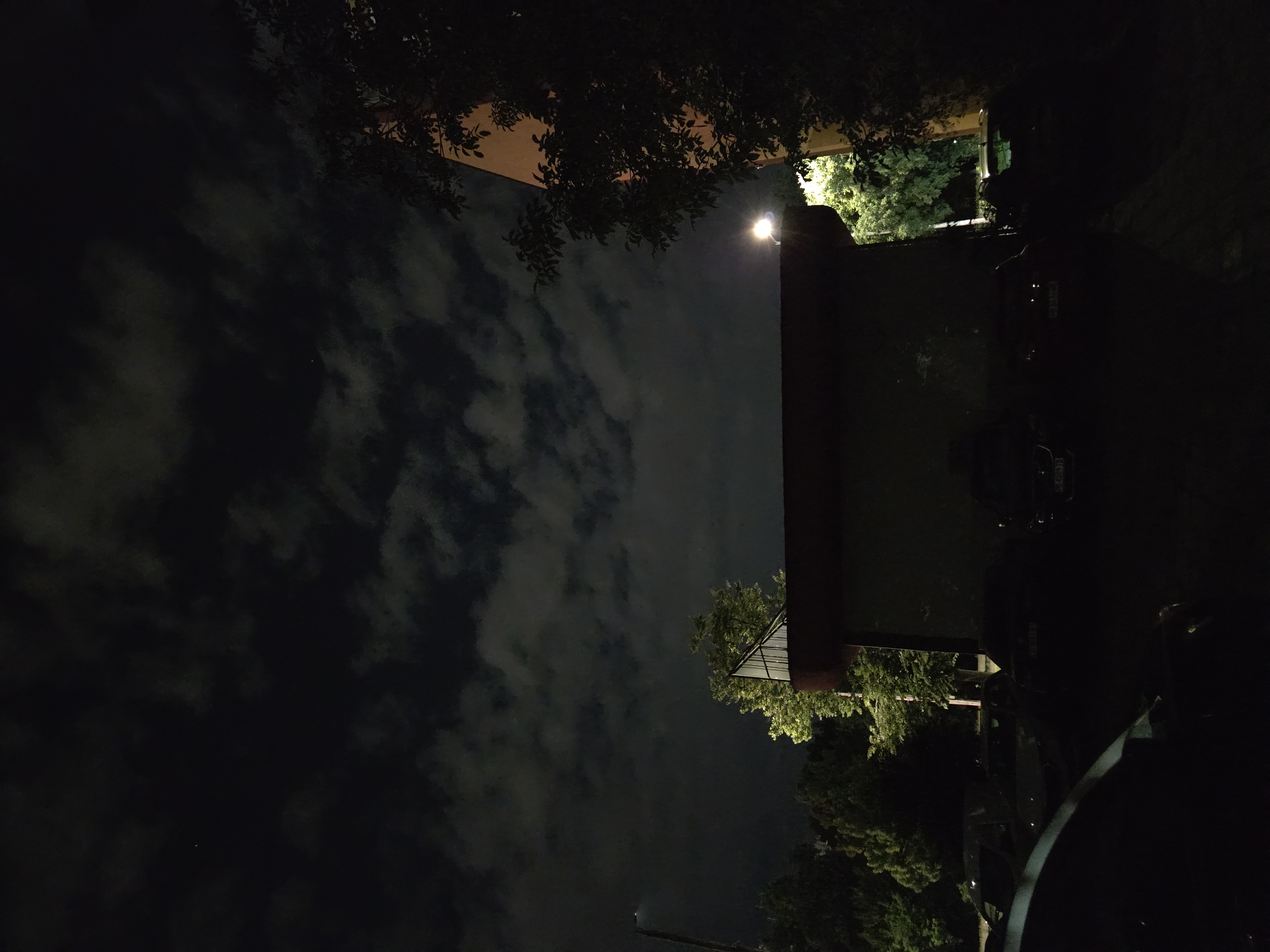 The image depicts a night scene. The sky is filled with dark clouds, illuminated by the moon or an artificial light source. In the foreground, there are trees and a building. The building appears to have a light on, and cars are parked nearby. The image's location is unknown.