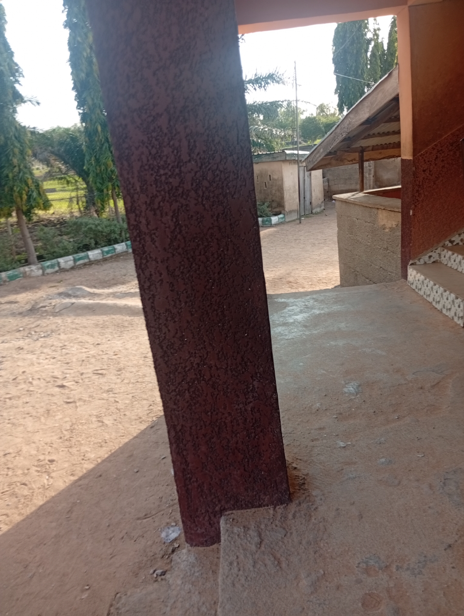 The image shows an outdoor scene with a prominent dark brown, textured vertical pillar in the foreground, obscuring a portion of the view. Beyond the pillar, there is a dirt ground surface with some patches of lighter sand and a clear shadow cast diagonally across it. To the left, a row of manicured green bushes and trees lines a dirt path, with some buildings visible in the background. One building appears to be a small, light-colored structure with a corrugated metal roof supported by wooden beams, possibly a market stall or shelter. To the right, a concrete structure with a tiled edge and steps is visible, suggesting a building entrance. Tall, slender trees are scattered throughout the background, and the sky is bright and overcast. The overall setting appears to be a public area in Jalingo, Nigeria, possibly near a school or community space, characterized by its dry, dusty environment and simple architecture. There are no people visible in the image.