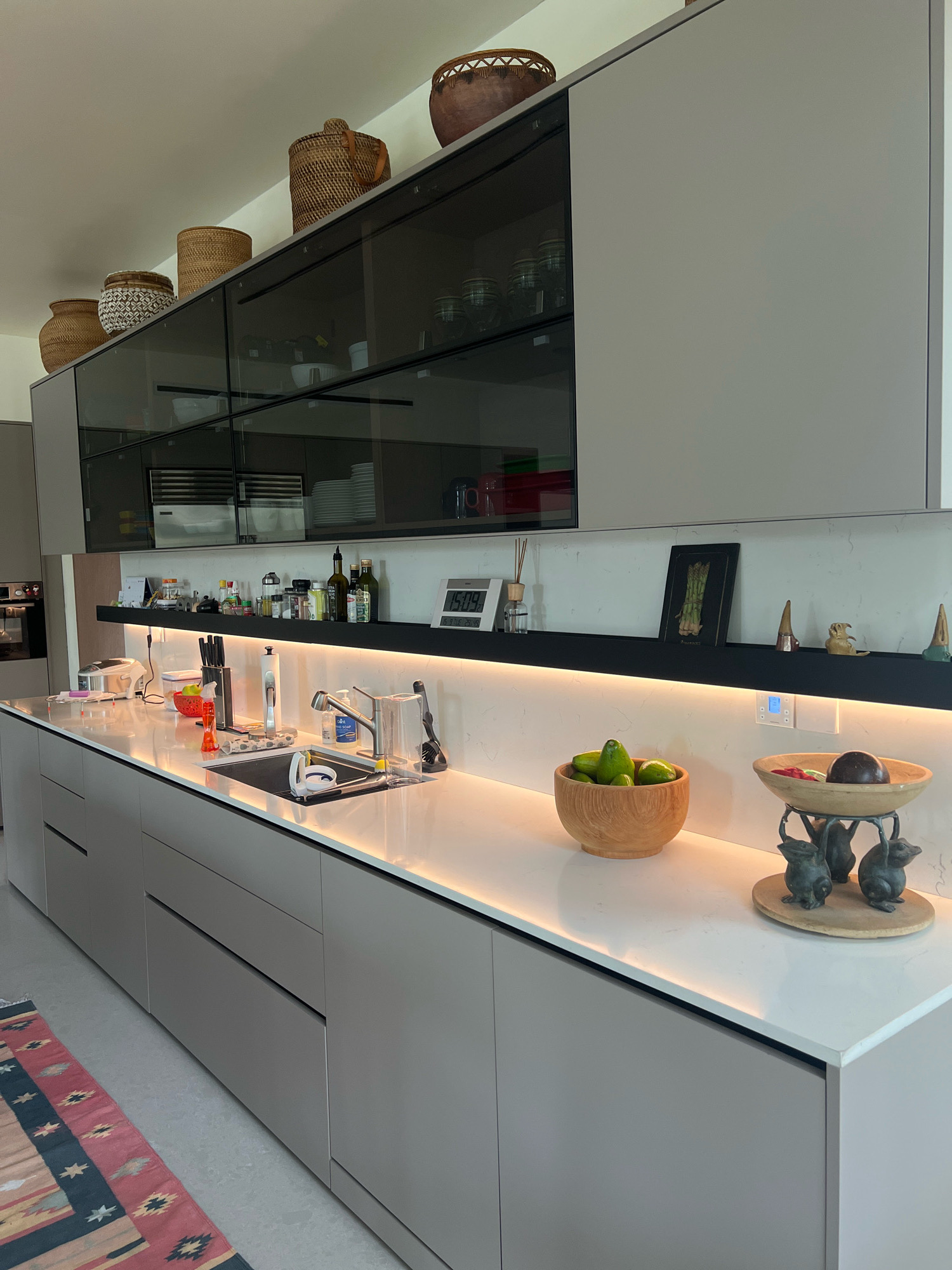 This is a forensic, matter-of-fact description of a modern kitchen scene in Ngaglik, Indonesia. The image focuses on a long, light-colored countertop that runs along a series of gray cabinetry. On the countertop, to the right of a stainless steel sink, there is a wooden bowl filled with green avocados, and to its right, a decorative stand holding a dark brown object and a few small figurines that appear to be animals. Behind these items, a black shelf is mounted on a textured white backsplash. This shelf holds several small decorative items, including a framed picture of asparagus, and various bottles and jars. Above this shelf, a set of dark glass-front cabinets are visible, with some baskets and earthenware vessels displayed on top of them. Integrated LED lighting illuminates the backsplash and the underside of the upper cabinets. To the left of the sink, the countertop holds a knife block, a roll of paper towels, a dish rack, and various cleaning supplies. Further left, a toaster and a pink object are visible. The cabinetry below the countertop appears to be handleless, with thin black lines indicating drawers and doors. A colorful patterned rug is partially visible on the floor to the left. The overall environment suggests a clean, well-organized, and contemporary kitchen space.