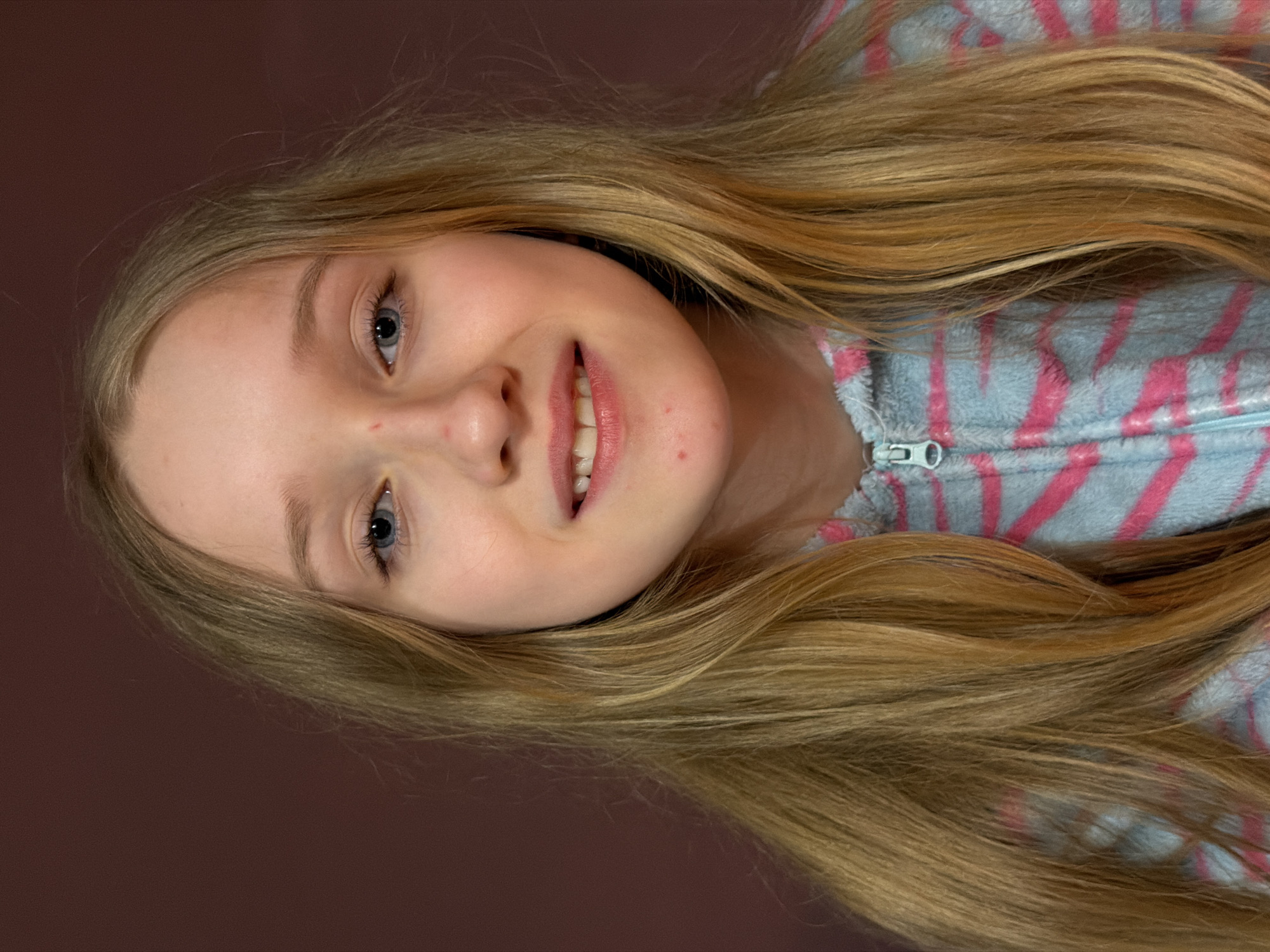 A portrait of a young Caucasian girl with long blonde hair. She is smiling slightly and wearing a zip-up hoodie with light blue and pink stripes. The background is a solid brown color. No evidence indicates the location.