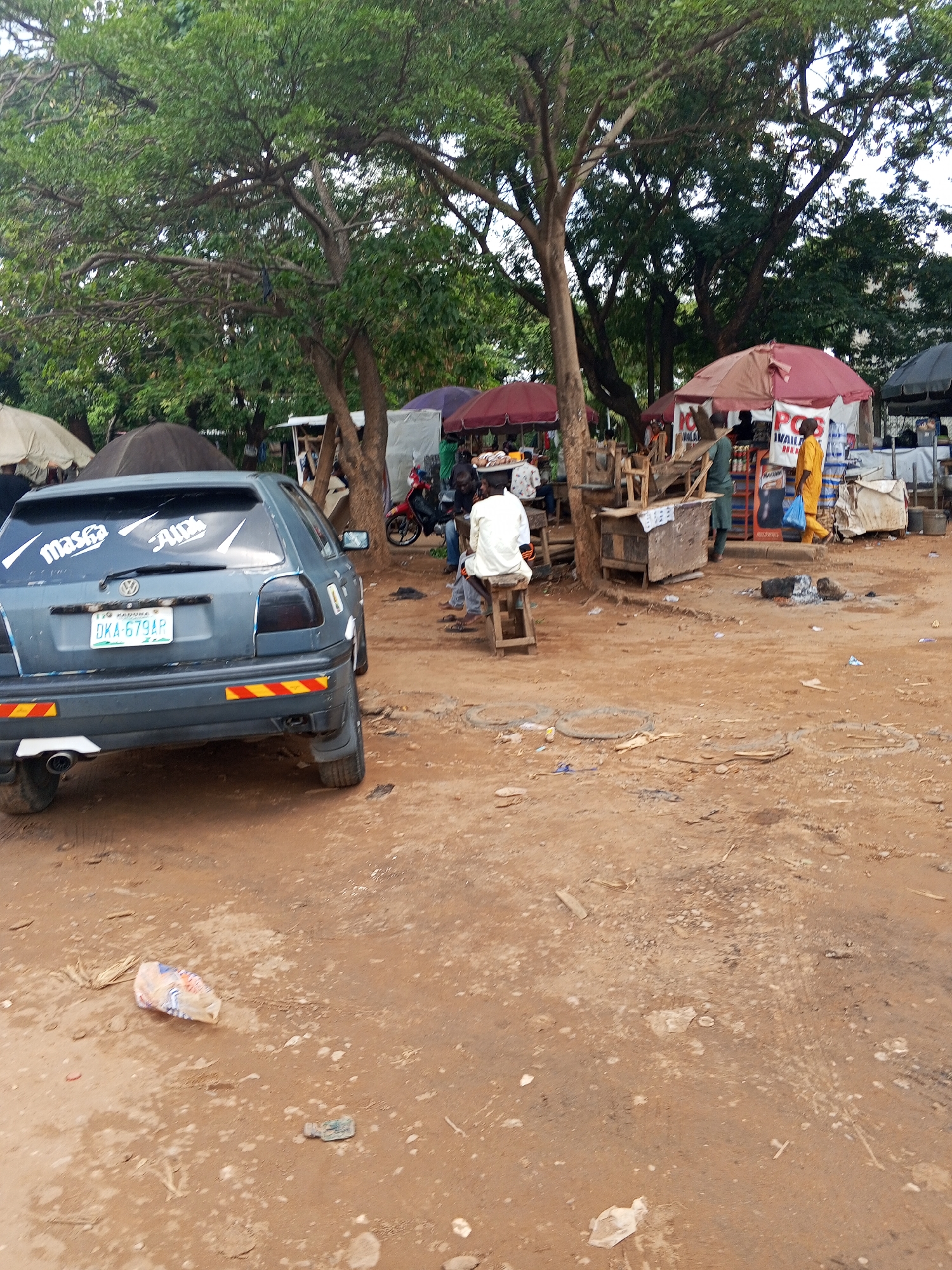 The image depicts an outdoor scene in Abuja, Nigeria, characterized by a dirt ground. A gray Volkswagen Golf is parked on the left, with a license plate reading "DKA-679AR." The car features a "masga" and "Aupri" sticker on its rear window. A man in a white shirt is sitting on a small stool, partially obscured by a tree trunk. In the background, there are several makeshift stalls and vendors under umbrellas. Some individuals are standing or walking around the stalls. There is debris and trash scattered on the ground, including a plastic bag. The lighting suggests it is daytime. Green trees provide some shade over the area.