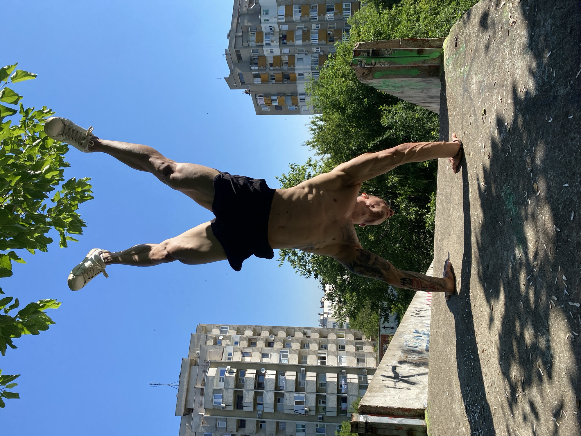 A shirtless man with tattoos on his arm performs a handstand on a concrete platform. He is wearing black shorts and white sneakers. His legs are extended upwards, and his body is balanced on his hands. In the background, there are apartment buildings and lush green trees, indicative of an urban setting. The sky is clear blue. The concrete platform has some graffiti on it and is littered with debris. The scene appears to be outdoors in a public area. The man is positioned in the foreground, with the city landscape behind him, suggesting the location is within Pleven, Bulgaria.