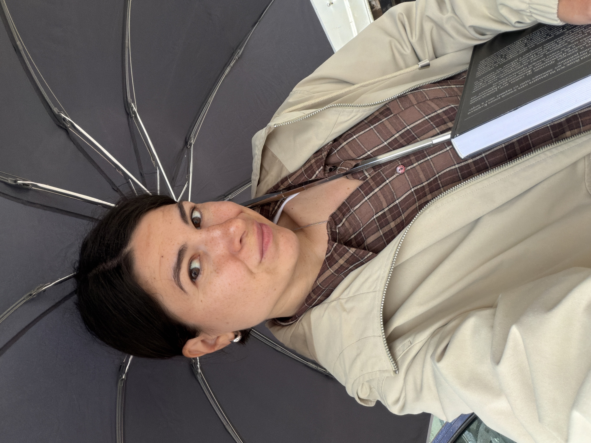 The image, rotated 90 degrees clockwise, presents a close-up of a woman under an open dark umbrella, holding a black hardcover book. The woman, appearing to be young to middle-aged, has dark hair parted on the side, and a face adorned with freckles. She looks directly at the camera with a neutral expression, eyes wide open.
She is dressed in a brown and cream plaid shirt layered under a light beige or cream zippered jacket. A silver chain necklace and small silver hoop earrings are visible. The dark grey or black umbrella canopy and its silver structural spokes occupy a significant portion of the frame, framing her face and torso. The book, held in her right hand, has white pages and a black cover with white text printed in Cyrillic script. Portions of the text visible include "рейтинге", "не имеет", "мест", "Терволда", and "миро", indicating Russian or a similar language. The background behind her is largely obscured by the umbrella, with only faint hints of an outdoor setting visible in the upper right. The scene appears to be set outdoors in Budva, Montenegro, likely on an overcast day or in a shaded spot given the use of the umbrella.