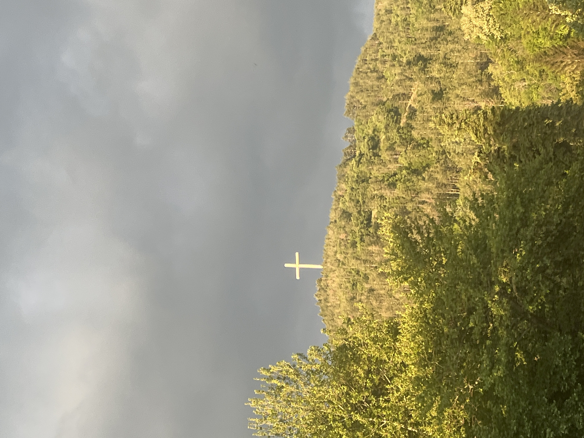A view of the Millennium Cross in Blagoevgrad, Bulgaria, with a dark, stormy sky in the background. The cross, a white structure, stands atop a hill covered with trees and vegetation. Part of the foreground is also filled with foliage.