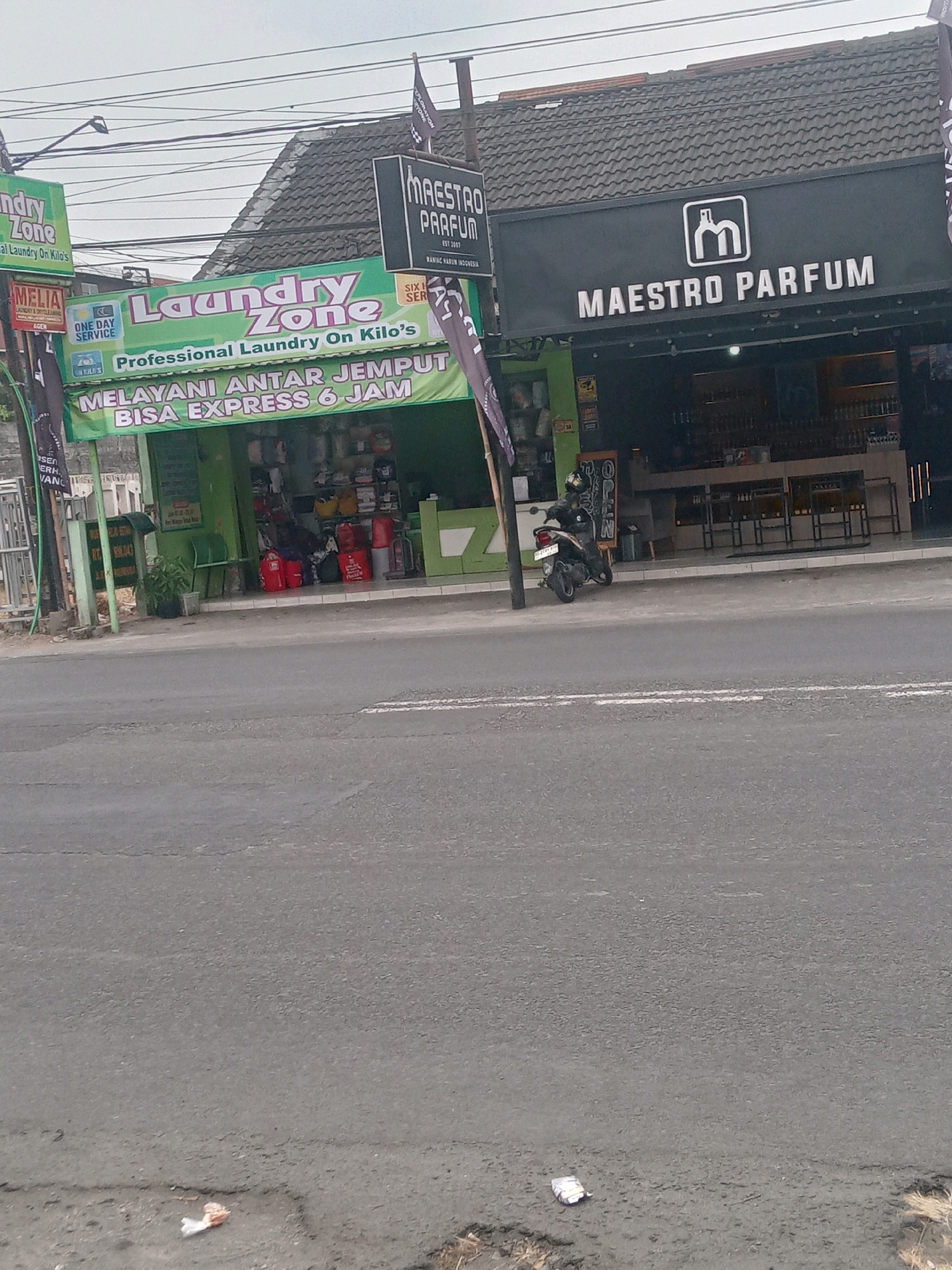The image shows a street scene in Maguwoharjo, Indonesia, with two storefronts prominently displayed. On the left is a bright green establishment with "Laundry Zone" written in large, colorful letters across its facade, along with smaller text stating "Professional Laundry On Kilo's" and "Melayani Antar Jemput Bisa Express 6 Jam" (Serving Pick-up and Delivery, Can be Express in 6 Hours). A smaller red sign with "MELIA" is visible above the laundry. To the right of the laundry, a dark gray storefront with "MAESTRO PARFUM" in large white lettering is visible, featuring a stylized "M" logo. The Maestro Parfum store appears to be a retail space with shelves stocked with bottles visible through its open front. The street in front of the shops is paved asphalt, with a white line marking a lane. A motorcycle is parked near the Maestro Parfum store, and a person wearing a helmet and dark clothing is seated on it. The buildings have tiled roofs. The sky above is overcast and light gray. Several power lines are strung across the top of the image.