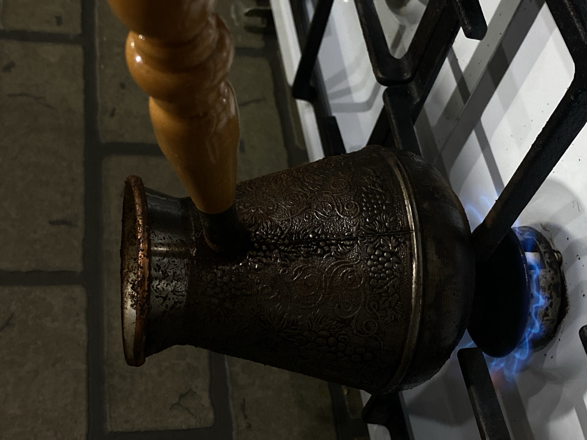 The image depicts an aged, dark metallic Cezve (Turkish coffee pot) with an embossed grape and vine pattern, positioned on a white gas stovetop. A light brown wooden handle extends from the right side of the pot. The Cezve rests on a black metal grate above a lit gas burner, from which a distinct blue flame is visible, actively heating the pot. The pot's rim shows wear and residue. The stovetop surface exhibits minor discoloration. The background consists of a dark, brick-patterned wall. The scene is dimly lit, with the flame providing the primary illumination. The location is in the city of Sochi, Russia. No individuals are present.