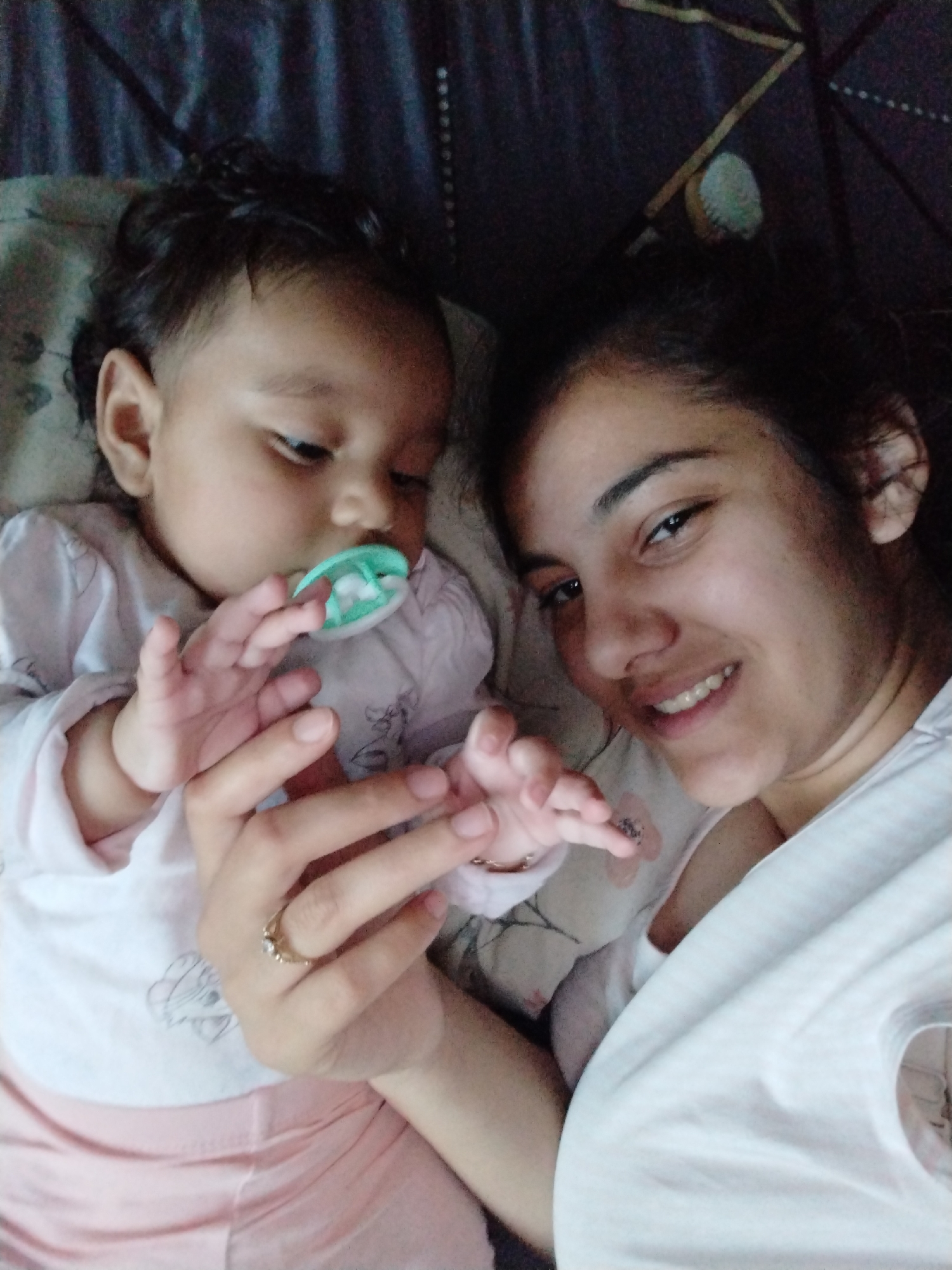 A medium-shot, slightly high-angle photograph shows a young woman and a baby lying next to each other on a patterned surface. The woman is on the right side of the frame and the baby is on the left. Both subjects are wearing light-colored clothing. The baby has a pacifier in its mouth and is holding the woman's hand. The woman is smiling at the camera. The surface they are lying on appears to be bedding with a floral or geometric pattern. The location is unknown.