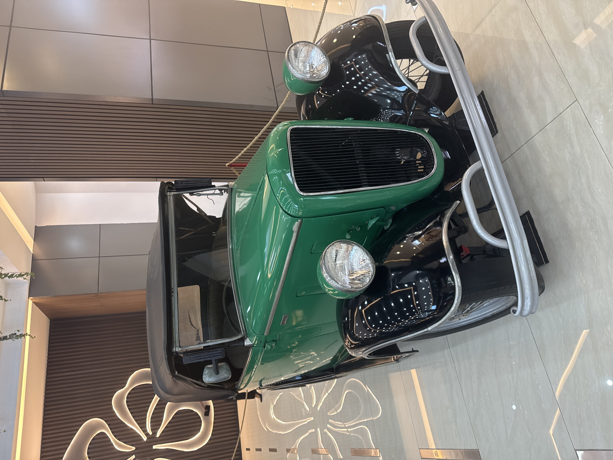 The image shows a vintage green and black car with a chrome bumper, front grill, and headlights. It is parked inside a building, likely a showroom or museum, as suggested by the modern tile flooring and decorative wall paneling behind the car. The location is Unknown, Unknown.