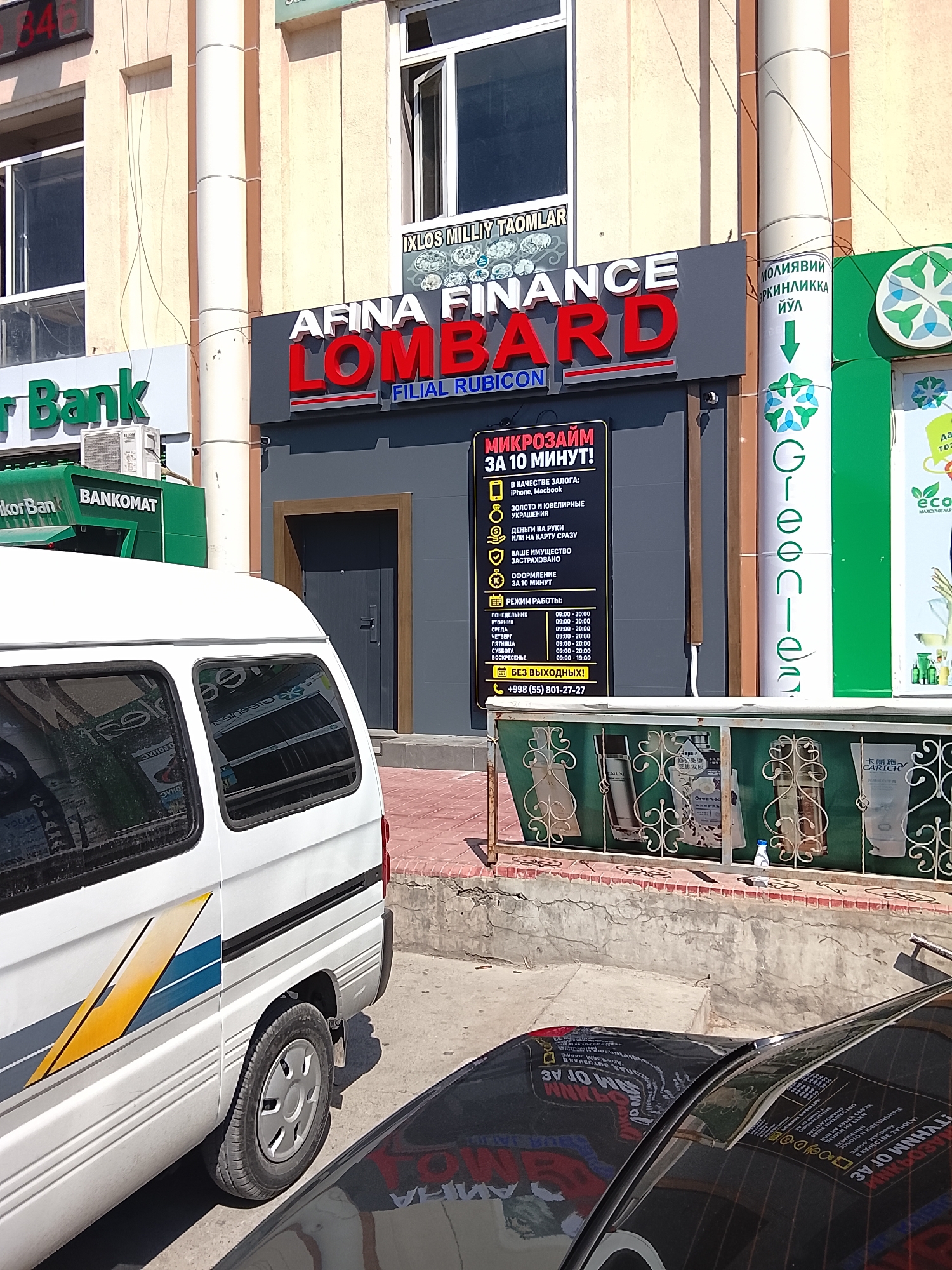The image shows a street scene in Fergana City, Uzbekistan, likely in an urban commercial area. In the foreground, the side of a white van with blue and yellow stripes is visible. To the right of the van, a black car's windshield reflects the surrounding buildings. Behind the van, a building features several storefronts. One prominent sign reads "AFINA FINANCE LOMBARD FILIAL RUBICON" in large red and white letters. Adjacent to it is a dark gray storefront with a closed gray door. To the left of the "AFINA FINANCE" sign, a green sign with white lettering reads "Bank" and below it "BANKOMAT." Further to the left, another sign with green lettering appears to be for a bank as well. Another building segment on the right has a green facade with a vertical white logo resembling a stylized snowflake or flower. A smaller sign in yellow and black provides details about "MIKROZAIM ZA 10 MINUT!" (Microloan in 10 minutes!) including services offered and operating hours. The ground is paved with concrete and some red brick. The overall environment suggests a commercial street with financial institutions and possibly a bank.