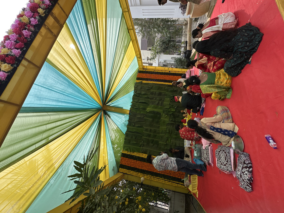 Under a colorful canopy of yellow, green, and blue fabric, people kneel on a red surface in what appears to be a cultural or religious ceremony. Floral decorations adorn the structure. Several individuals wear traditional attire, and objects like bags and plates are scattered nearby. The scene is captured at ground level, with trees and a building in the background.