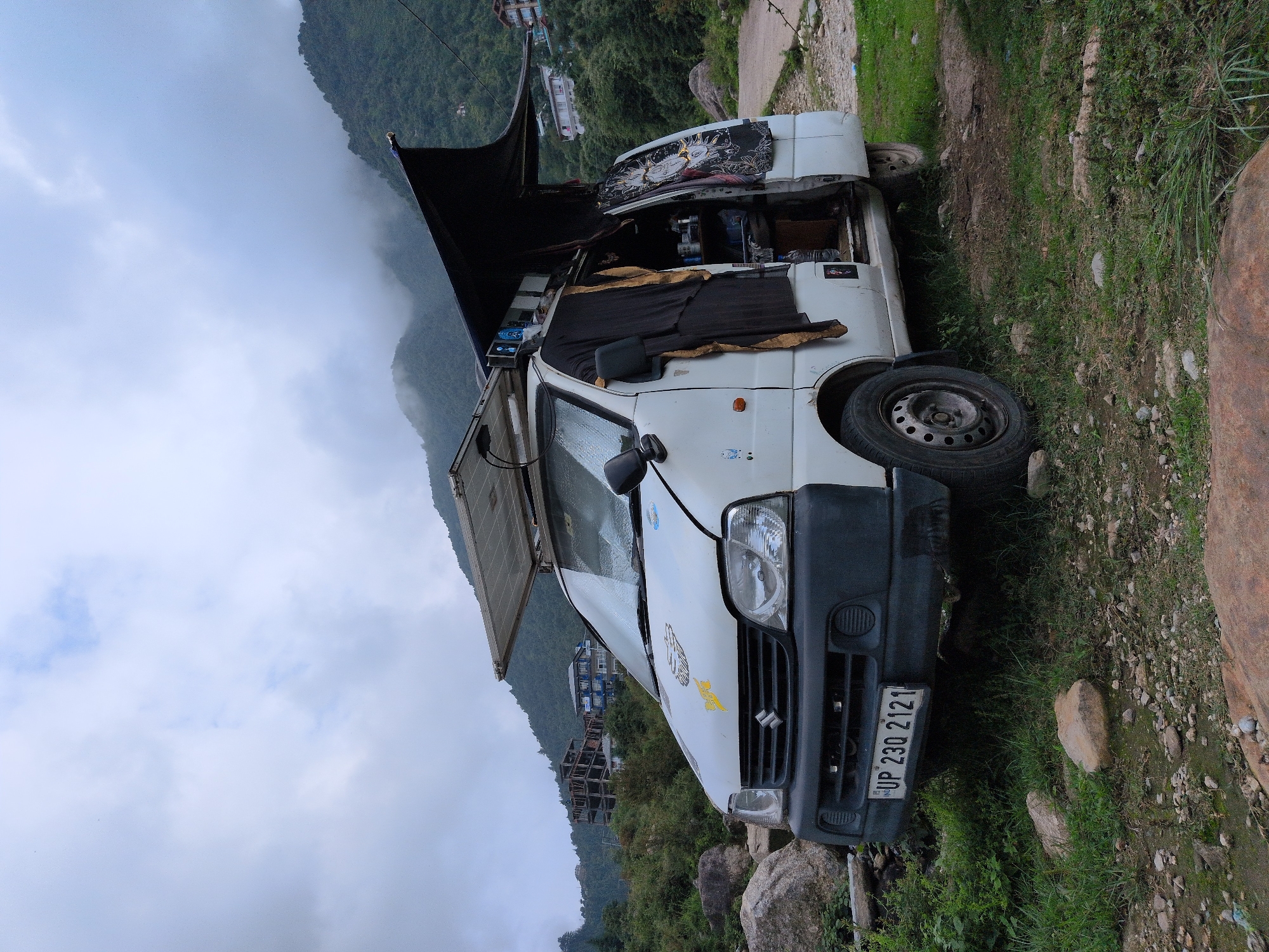 A white van, adapted for camping or living, is parked outdoors with a mountainous, forested landscape in the background. The van has solar panels mounted on its roof and an awning extended on the side. The driver's side door is open, revealing interior storage and items. A license plate reading "UP 230 2121" is visible on the front. The van is situated on uneven, grassy terrain with rocks and boulders. In the background, partially constructed buildings and houses are visible on the slopes of the mountains, suggesting a semi-developed area. The sky is overcast with clouds. The location is identified as Haini, India. There are no people or animals visible in the frame.